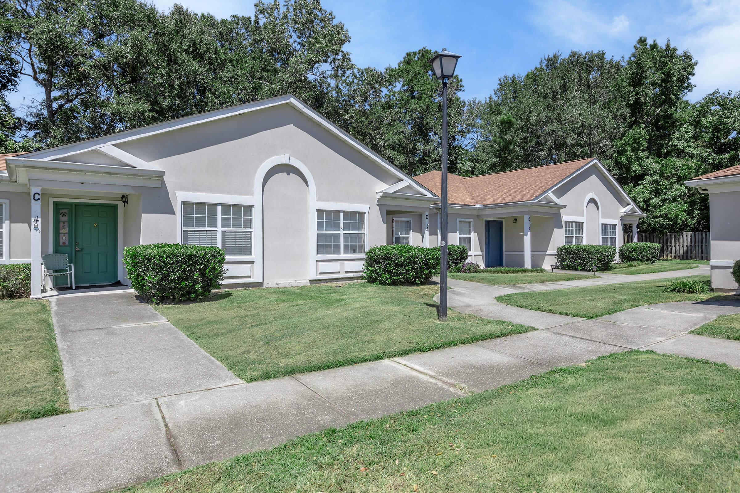 View of a residential complex featuring two single-story buildings with green doors. The well-maintained lawn is bordered by neatly trimmed bushes, and a lamppost stands nearby. Clear blue sky and trees in the background create a pleasant ambiance. Pathways connect the doors, enhancing the inviting atmosphere.