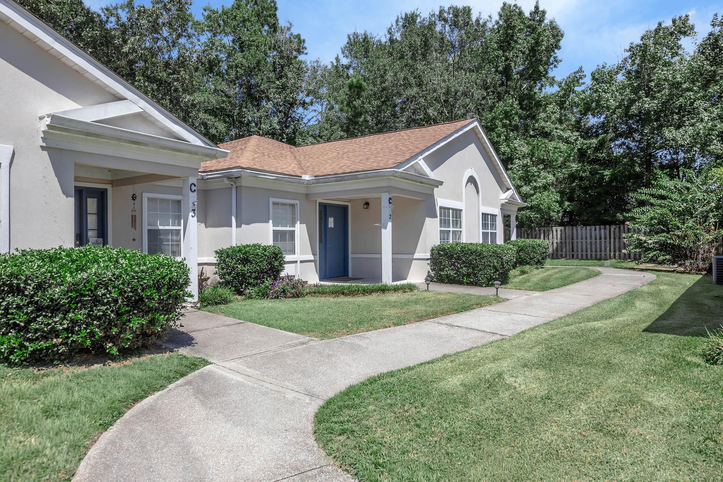 A well-maintained exterior of a residential property featuring a light-colored building with a brown roof. Neat landscaping includes trimmed bushes and a curved sidewalk leading to the entrance. Green grass surrounds the walkway, and lush trees are visible in the background under a clear blue sky.