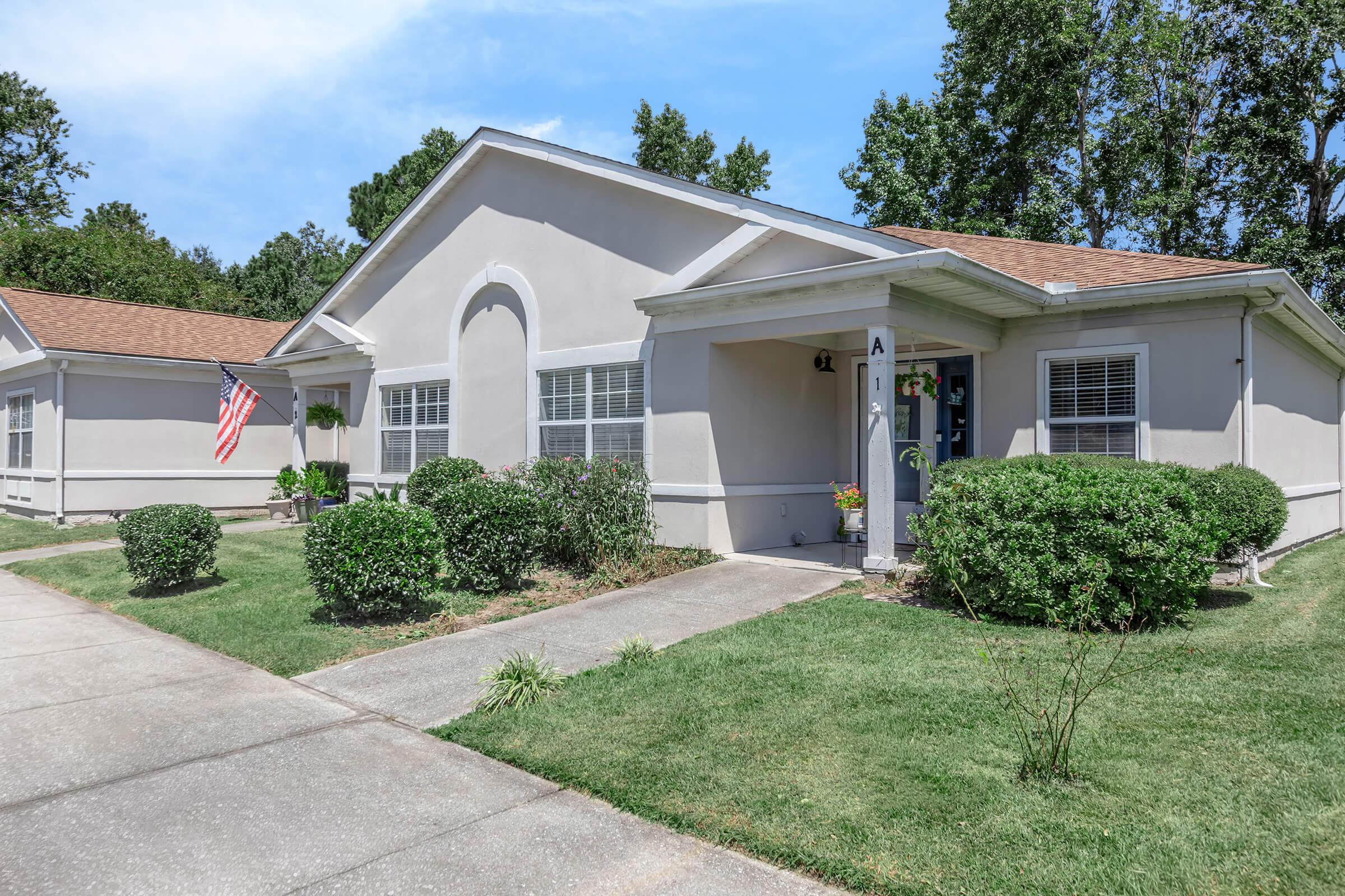 A well-maintained single-story house with a light gray exterior and a brown shingled roof. The entrance features an American flag and potted plants. The yard is landscaped with neatly trimmed bushes and grass. A paved walkway leads up to the door, and trees border the background under a clear blue sky.