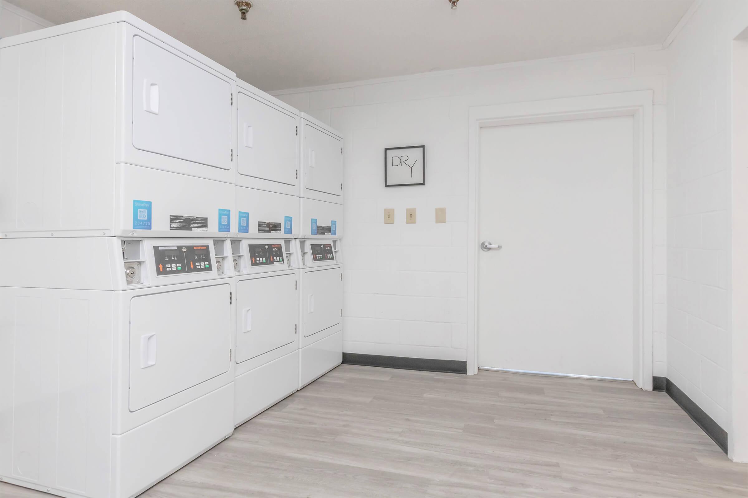 A clean and modern laundry room featuring multiple stacked washing machines and dryers against a white wall. The floor is light-colored, and there is a framed sign reading "DRY" on the wall beside a closed door. The overall ambiance is bright and tidy, suggesting a functional space for laundry activities.