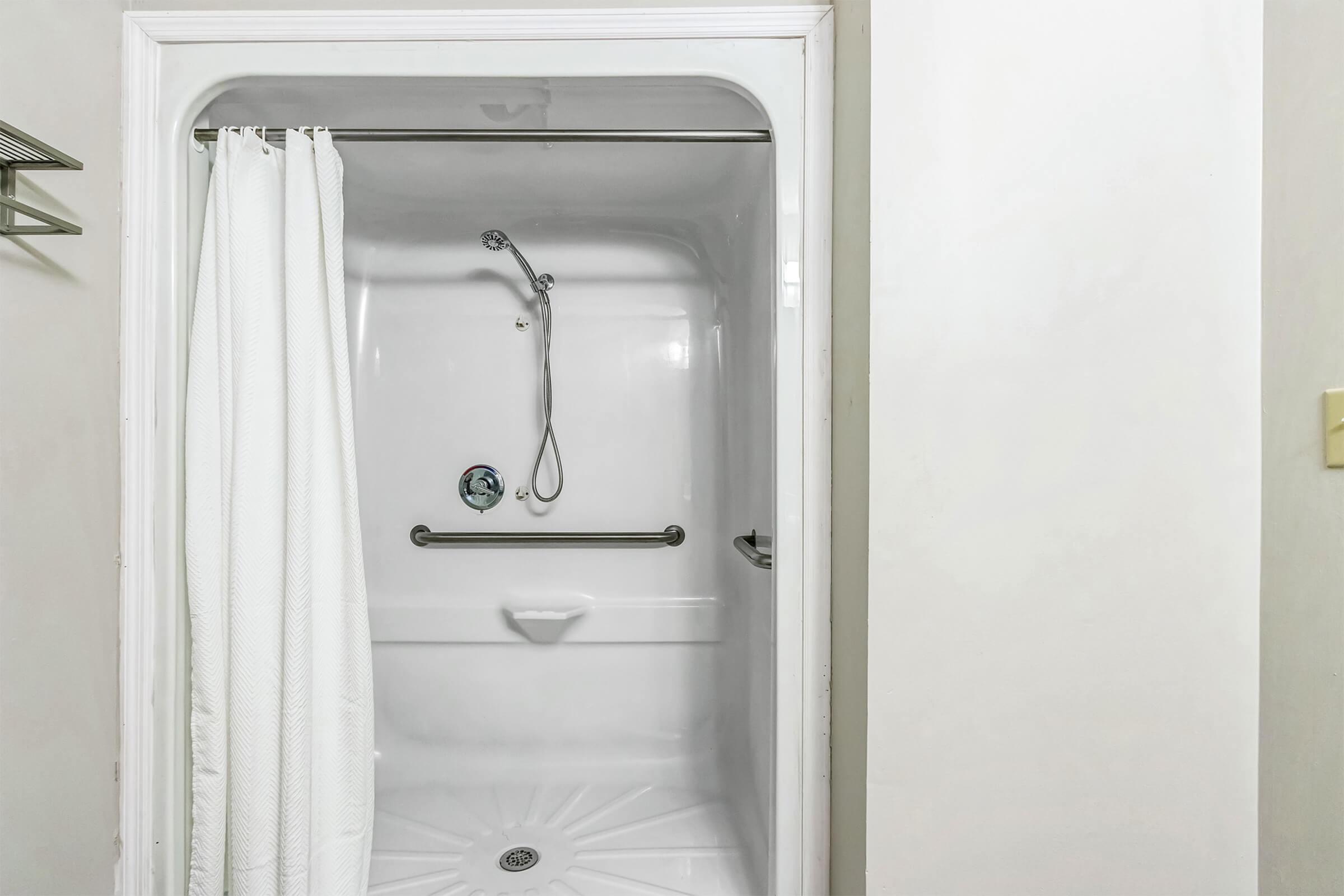 A clean, white shower stall featuring a grab bar, showerhead, and a curtain. The walls are smooth and light-colored, with a drain located at the center of the floor. The overall design is simple and functional, suitable for accessibility.