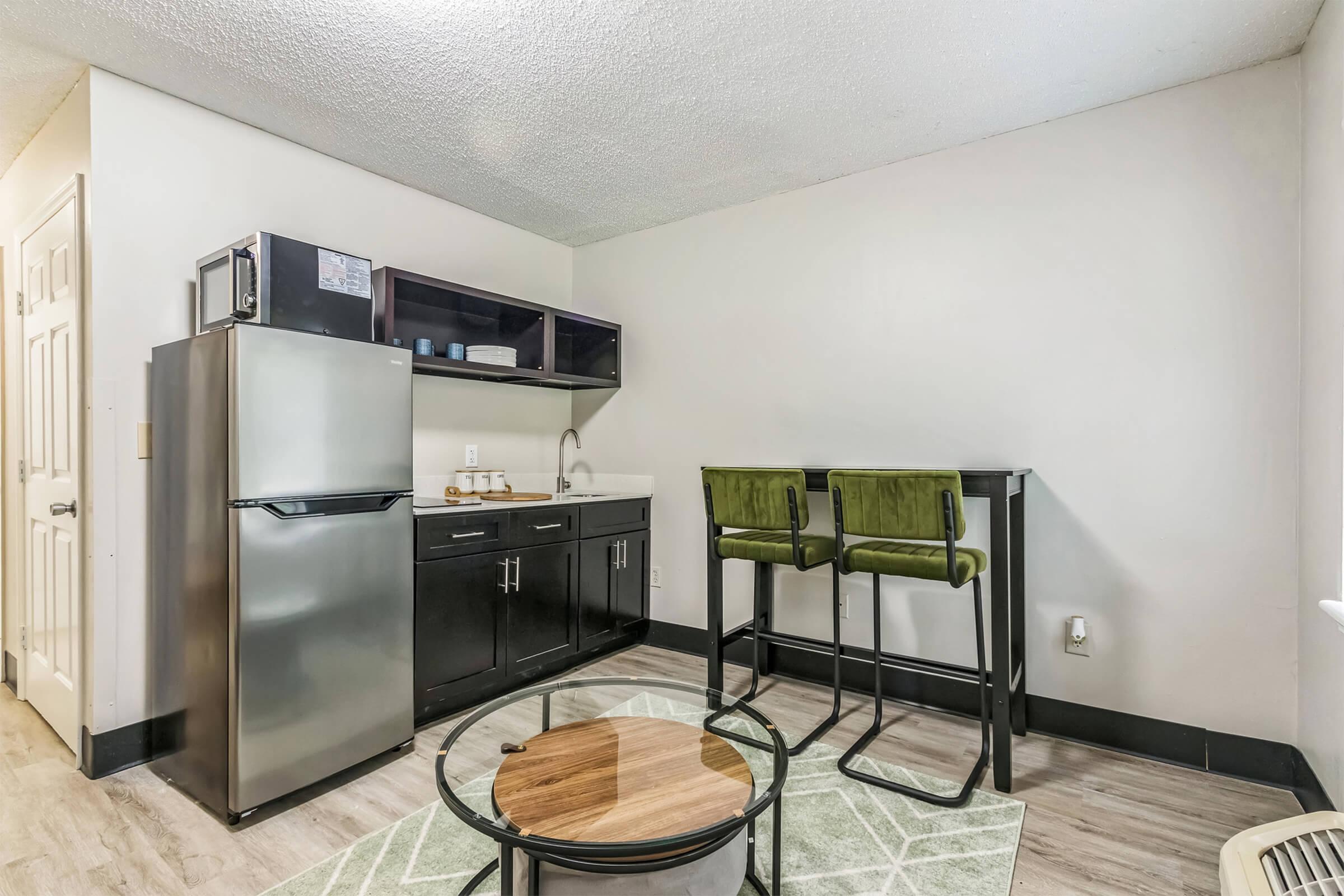 A compact kitchen area features a stainless steel refrigerator, a sink, and a countertop with modern storage above. Two stylish green barstools are placed at a matching table, alongside a circular wooden coffee table on a light-patterned rug. The walls are painted a neutral tone, enhancing the space's modern feel.