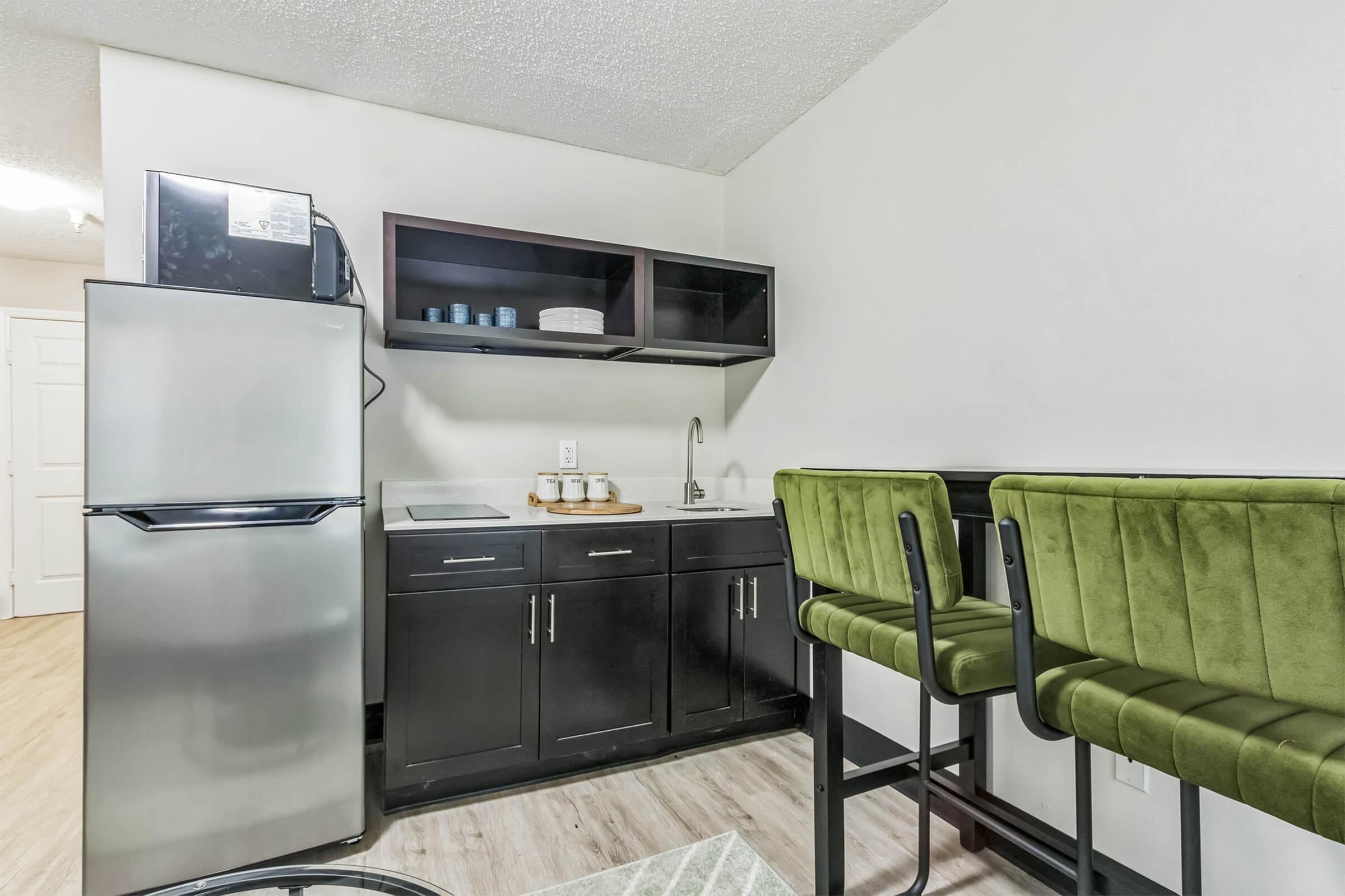 A modern kitchenette featuring a stainless steel refrigerator, a sink, and black cabinetry. Above the counter are shelves holding dishes and glassware. Two green bar stools with a plush texture are positioned at the counter, complemented by a light wood floor and a small area rug.