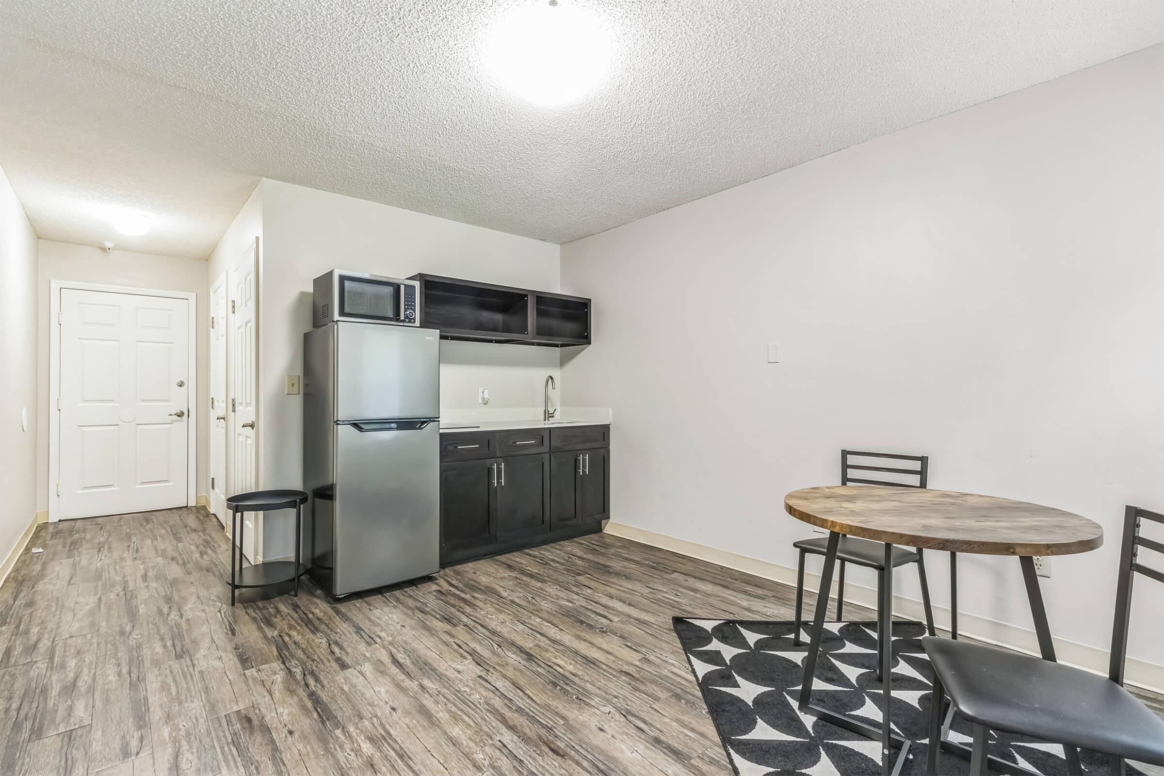 A small kitchen area featuring a refrigerator, sink, and modern cabinetry. A wooden round dining table with black chairs is positioned nearby. The floor is made of wooden planks, and the walls are painted in a light color. Ceiling lights provide adequate illumination in the space.