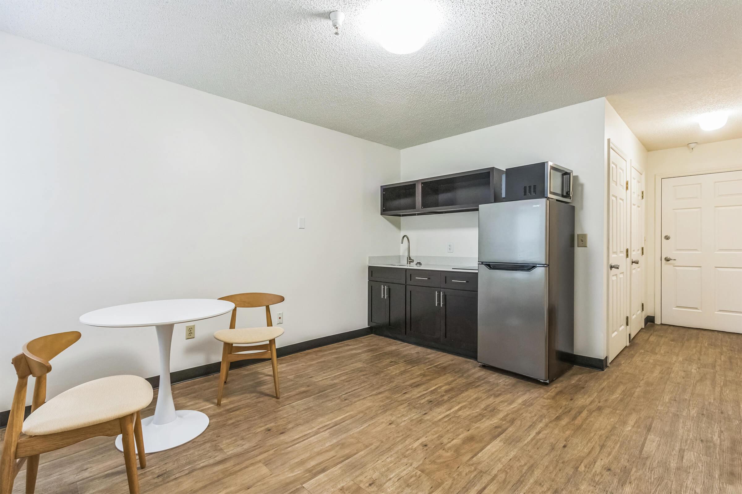 A modern, minimalist kitchenette area featuring a small round dining table with two wooden chairs, a stainless steel refrigerator, and dark cabinetry. The walls are light-colored, and the flooring is a wood-like laminate. A door leads out of the room, and there is overhead lighting in the space.