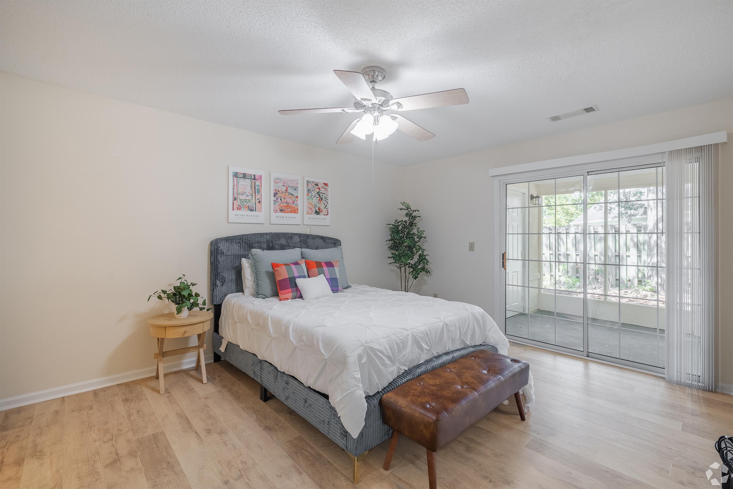 A bright and spacious bedroom featuring a large bed with a gray headboard and colorful pillows, a bedside table with a small plant, and a brown ottoman at the foot of the bed. A sliding glass door leads to a screened porch, and three framed art pieces are hung on the wall. Natural light fills the room.