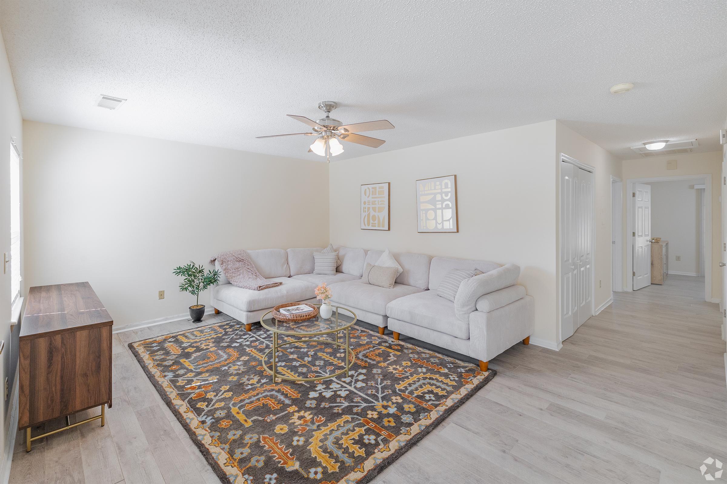 A bright and spacious living room featuring a light grey sectional sofa, a decorative area rug with intricate patterns, a wooden TV stand, and a small potted plant. The walls are painted in a soft shade, and there are framed art pieces on display. Natural light comes in through the windows.