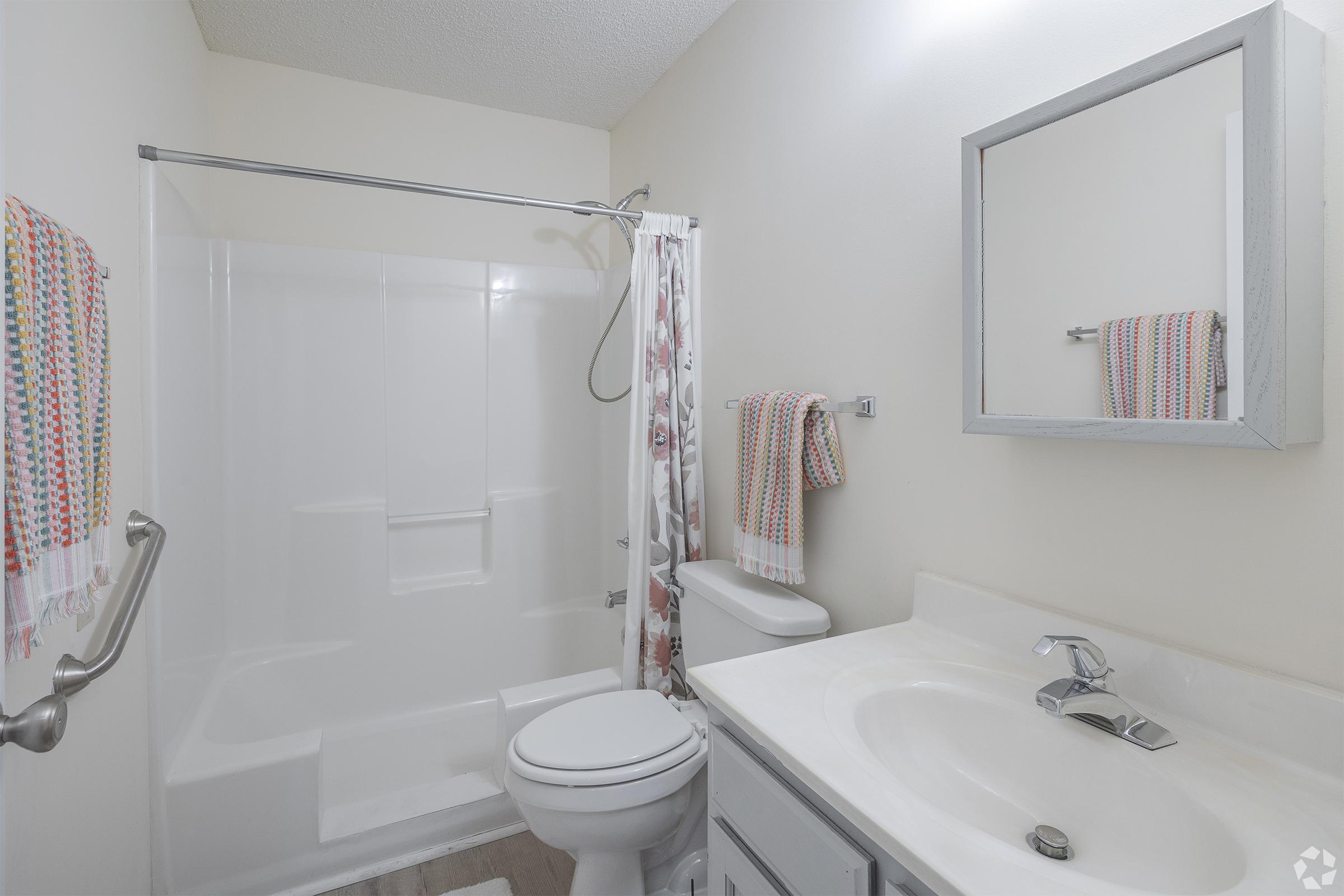 A clean, modern bathroom featuring a white shower with a clear curtain, a toilet, and a vanity with a sink. The wall is painted a light color, and there are colorful towels hanging on a rack. A small mirror above the sink reflects the room's simple and functional design.