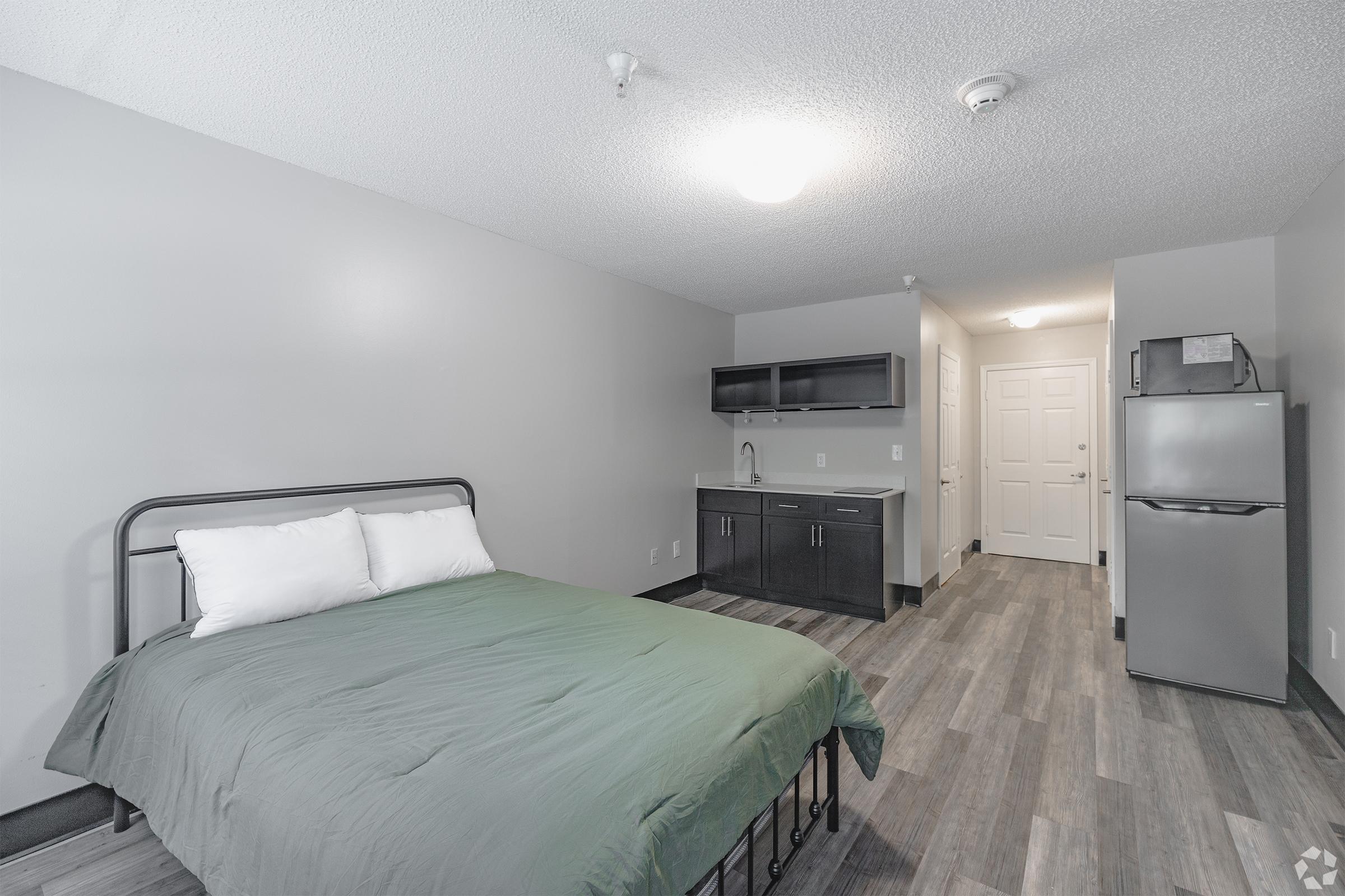 A simple, modern hotel room featuring a bed with a green blanket, a gray wall, and a kitchen area with dark cabinetry. A small refrigerator is visible next to the kitchenette. Bright lighting enhances the clean and minimalist design. The floor is covered with laminate in a wood-like finish.