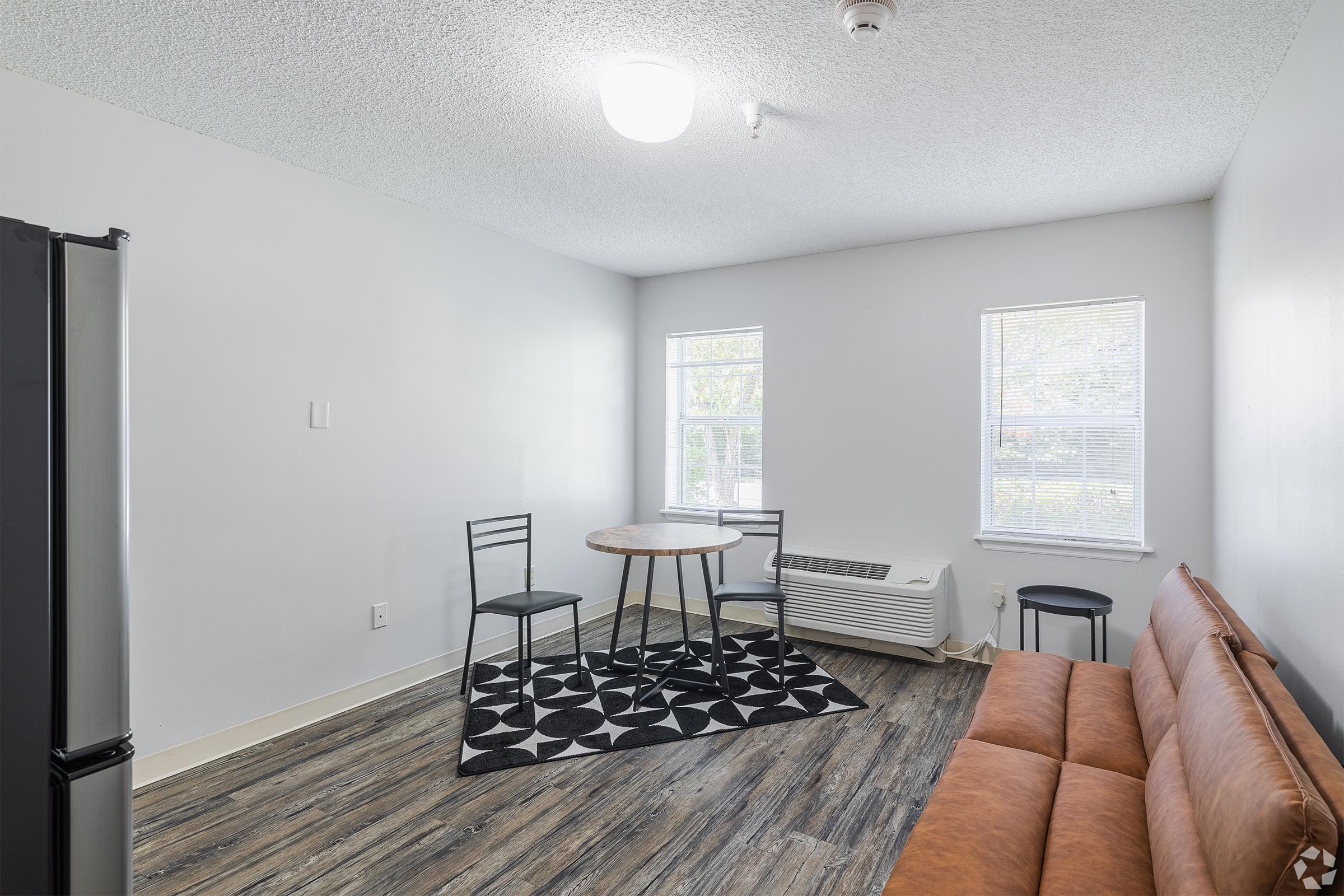A simple and modern interior of a small living space. Features a brown sofa, a round dining table with three chairs, a patterned black and white rug, and a window providing natural light. There is also a refrigerator in the corner. The walls are light-colored and the flooring has a wood-like appearance.