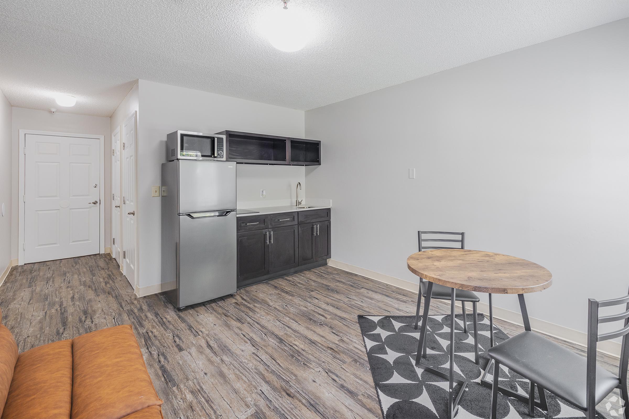 A modern kitchen and dining area featuring a gray refrigerator, black cabinets, a small round wooden table with two chairs, and a brown sofa in the foreground. The floor is wood-like with a patterned rug. The walls are painted light gray, creating a clean and contemporary look.