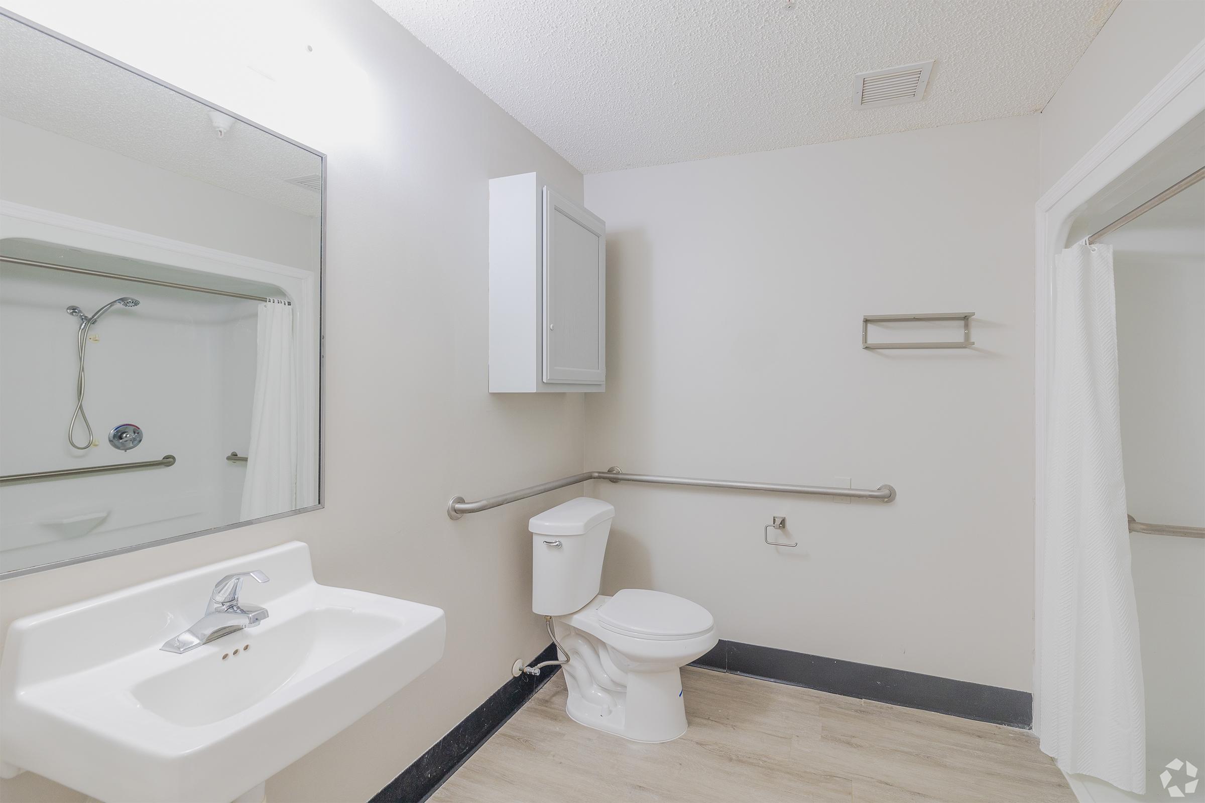 A simple, accessible bathroom featuring a white sink, a toilet, and a grab bar. There is a shower stall with a curtain, a mirror above the sink, and a cabinet on the wall. The walls are painted light gray, and the flooring is a light-colored laminate.