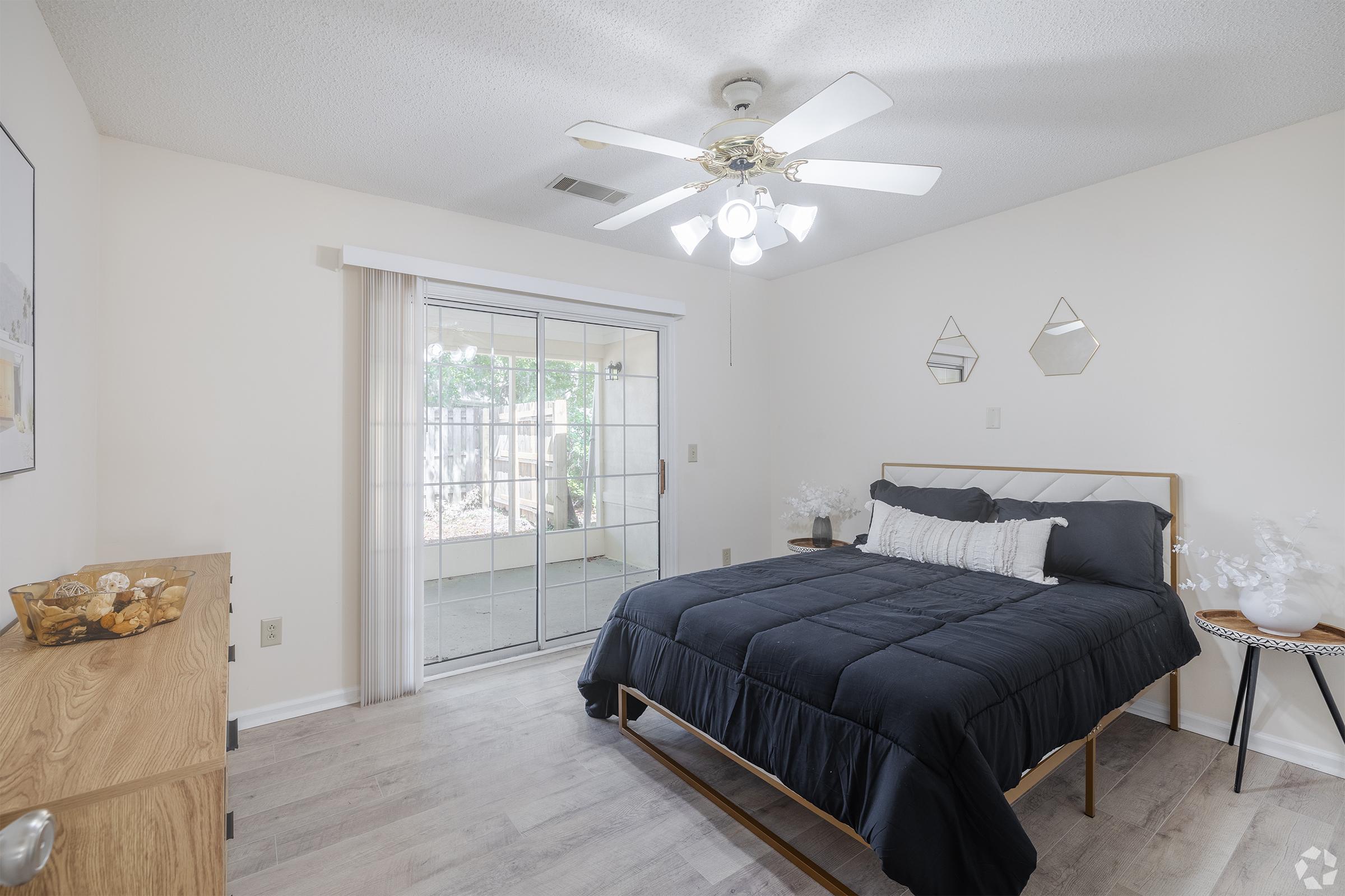 A cozy bedroom with a queen-sized bed covered in a dark blue comforter. The room features a ceiling fan, a sliding glass door leading to a patio, and decorative mirrors on the wall. Light-colored wood flooring and minimalistic furniture create a spacious and serene atmosphere.