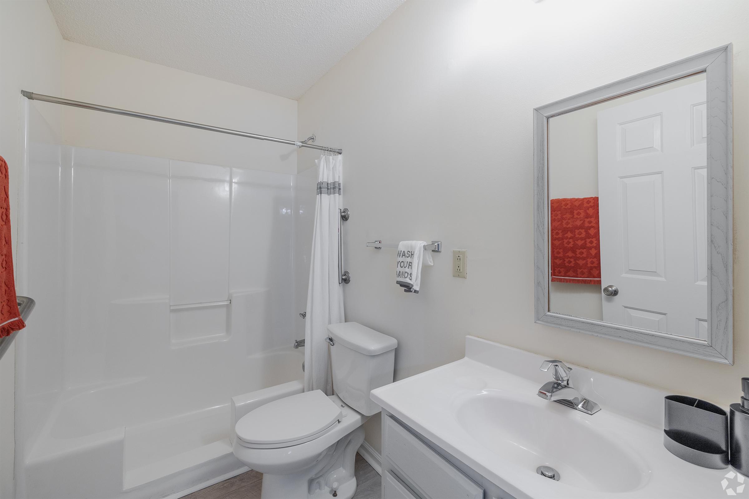 A clean and modern bathroom featuring a white shower enclosure with glass doors, a toilet, and a vanity with a sink and mirror. White walls and light-colored flooring create a bright atmosphere. Towels are neatly hung, and there are decorative elements, including a red towel and a sign on the towel rack.