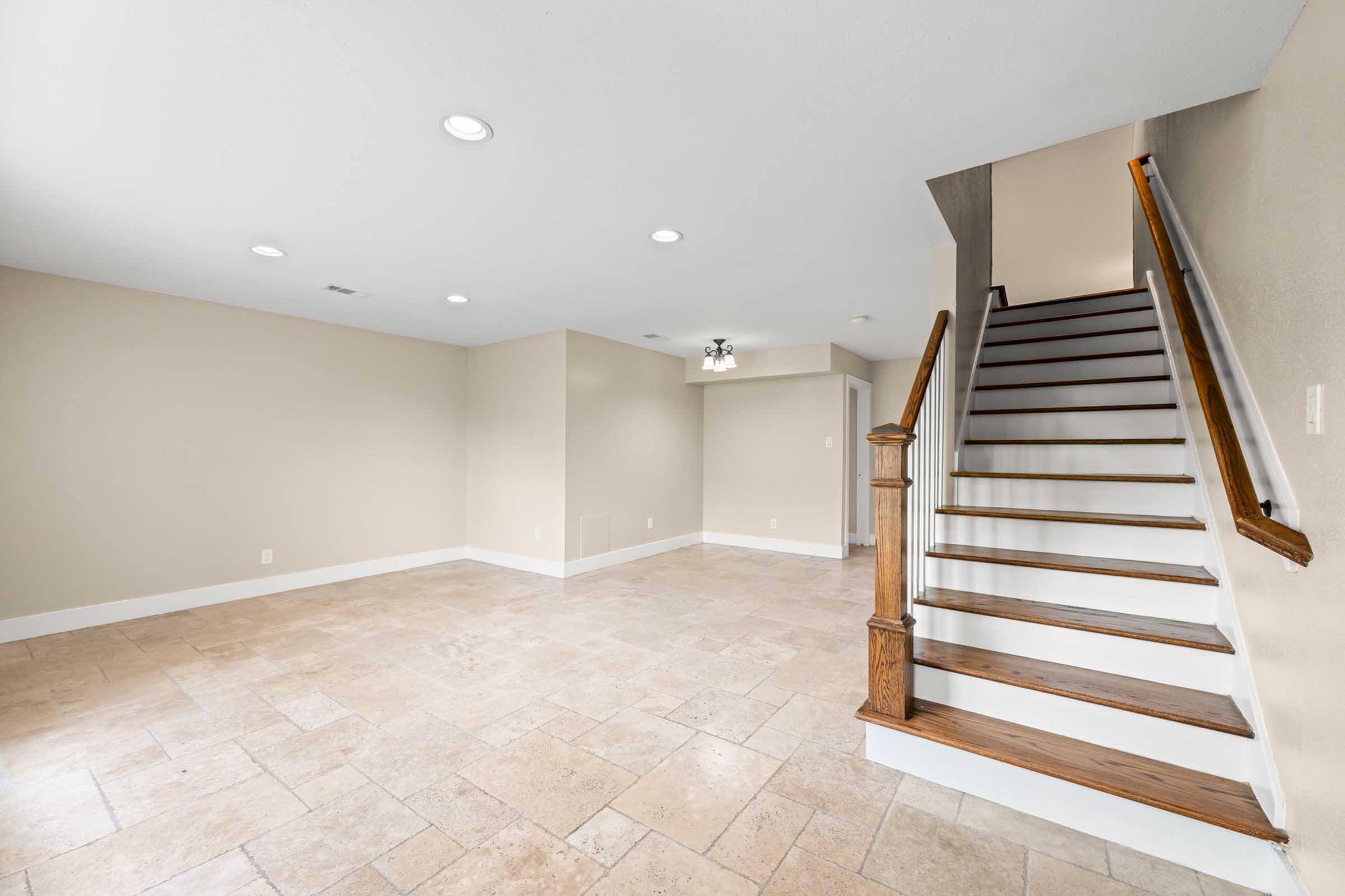 A spacious, empty room with beige walls and tiled flooring. There are several recessed lights on the ceiling, and a staircase with wooden railing leading to an upper level. The area is well-lit and has a clean, modern look, suitable for various uses.