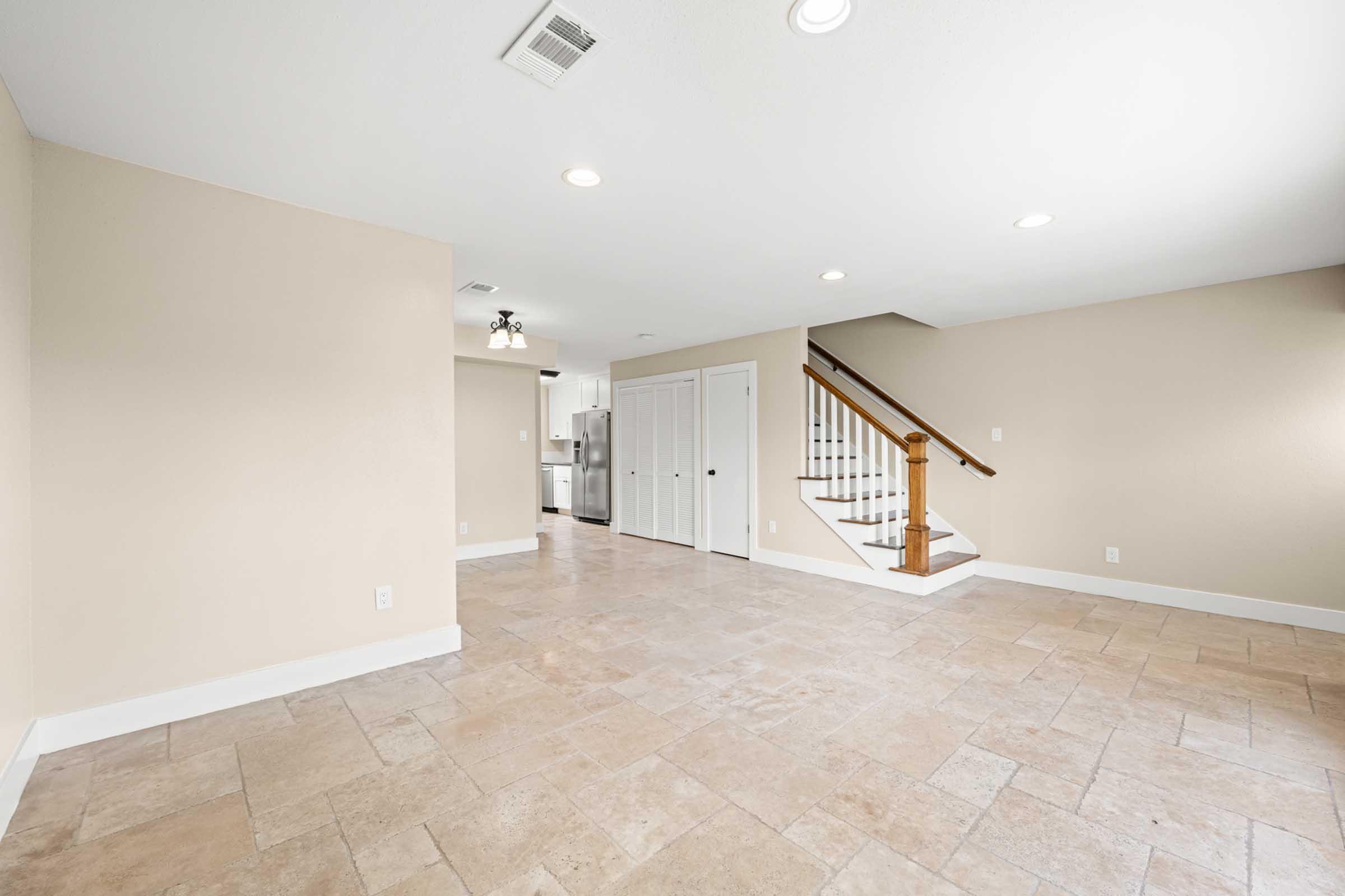 Spacious, empty interior room with beige walls and tile flooring. There is natural light from multiple recessed lights and a nearby staircase leading to an upper level. A doorway with white double doors is visible on one side, leading to another area of the home. The kitchen can be seen in the background.