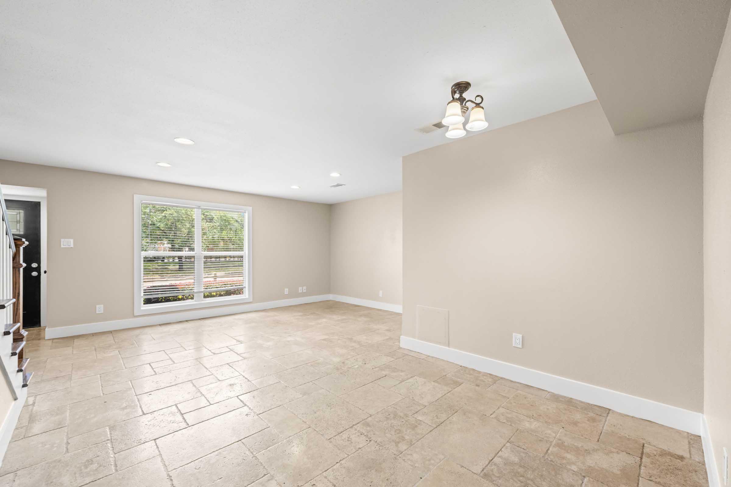 A spacious, empty room with beige walls and large windows allowing natural light. The flooring is made of light-colored tiles, and there are recessed lights in the ceiling. A light fixture is mounted near the center, and one wall has built-in shelves. The room is clean and modern, suitable for various uses.