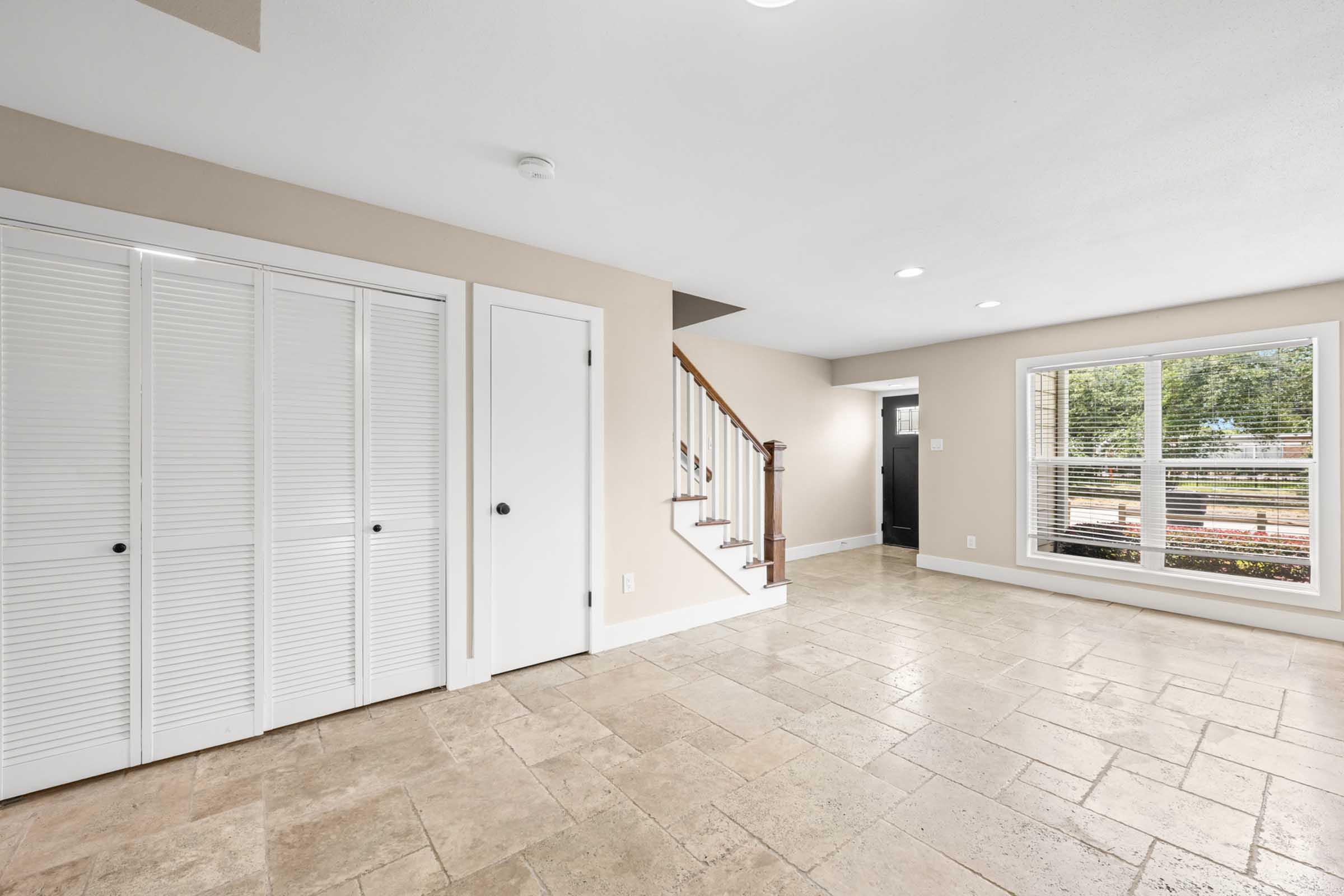 A spacious, well-lit interior room featuring beige walls, tile flooring, and natural light from large windows. There are built-in white closets on the left, a staircase leading to an upper level, and a doorway visible in the background. The overall ambiance is clean and modern.