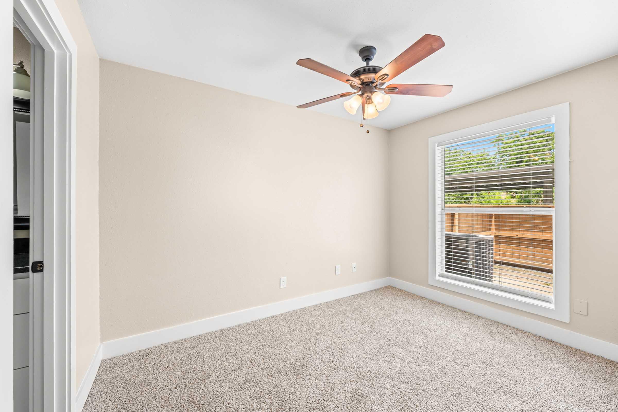 A well-lit, empty room featuring a ceiling fan with wooden blades, beige walls, and light-colored carpet. A window with blinds offers outdoor views, and there’s a door leading to another area in the background. The overall space appears clean and inviting, suitable for various uses.