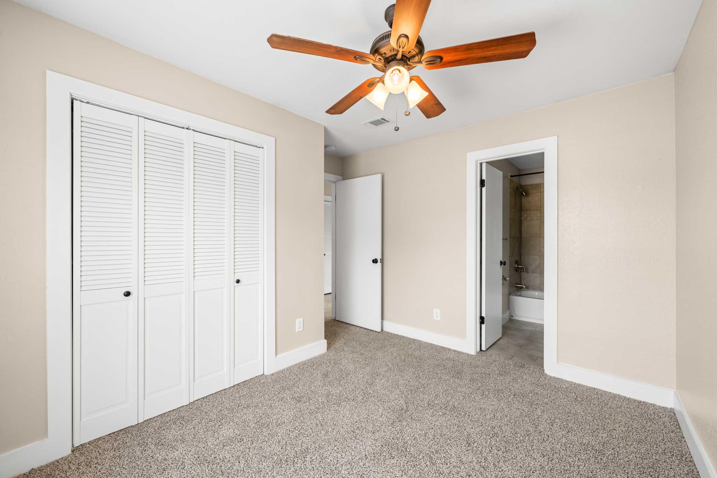 A well-lit room featuring beige walls, a ceiling fan with wooden blades, and plush carpet. The space includes a set of double doors with louvered panels, leading to a closet. A bathroom door is visible on the right, and natural light filters in, creating a warm and inviting atmosphere.