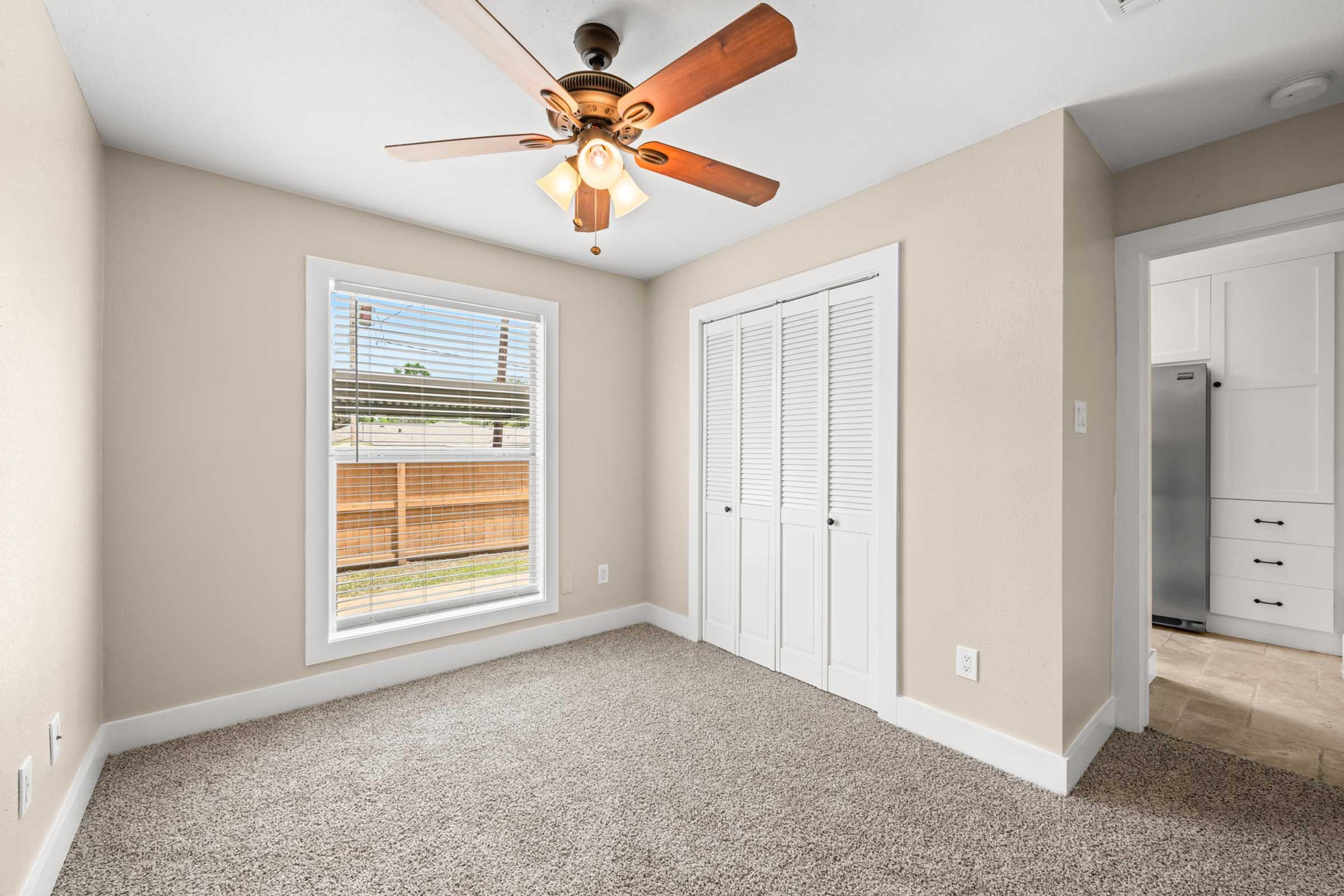 A well-lit bedroom featuring beige walls and a light brown ceiling fan. The room has a large window with blinds, offering a view of an outdoor space. To one side, there is a double closet with white doors. The flooring is carpeted in a light color, creating a cozy atmosphere.