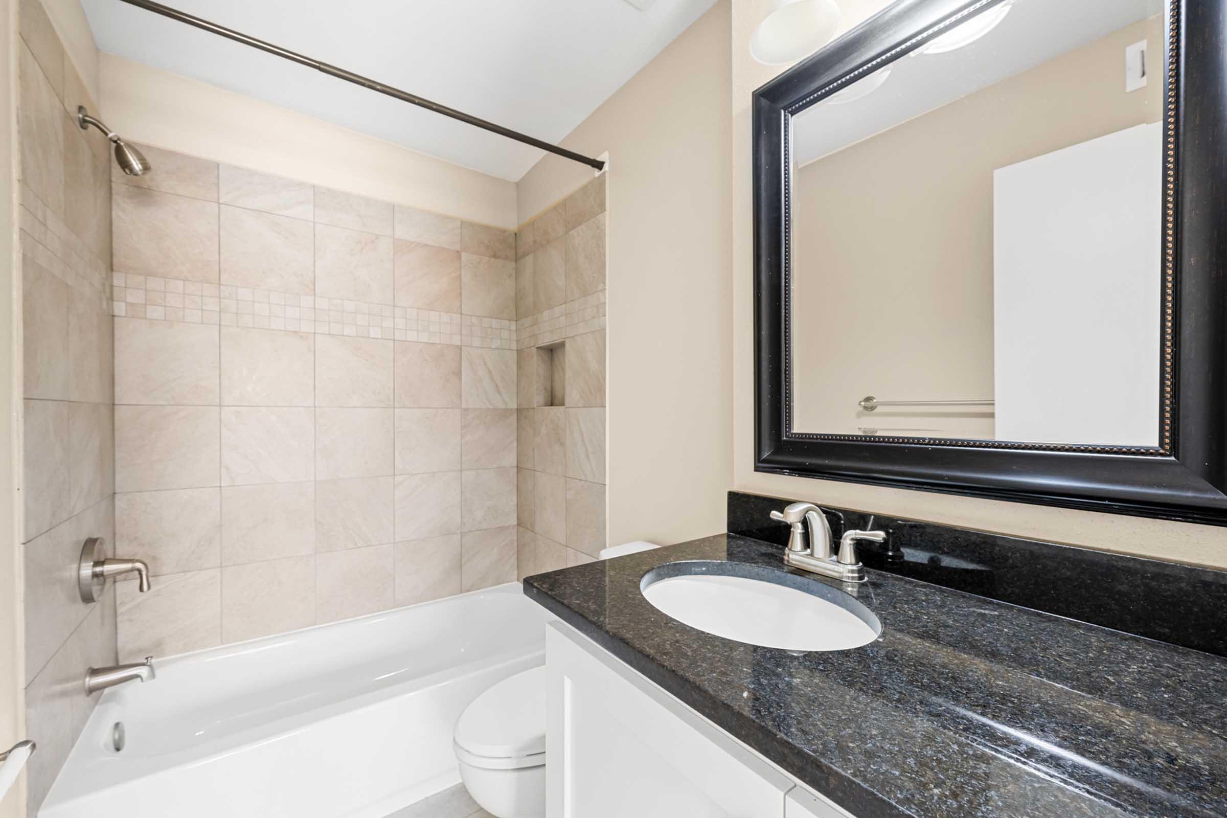 A modern bathroom featuring a shower-tub combo with beige tiled walls, a black granite countertop with an undermount sink, and a large rectangular mirror above the sink. The bathroom includes a white toilet and minimalistic decor, set against neutral wall colors.