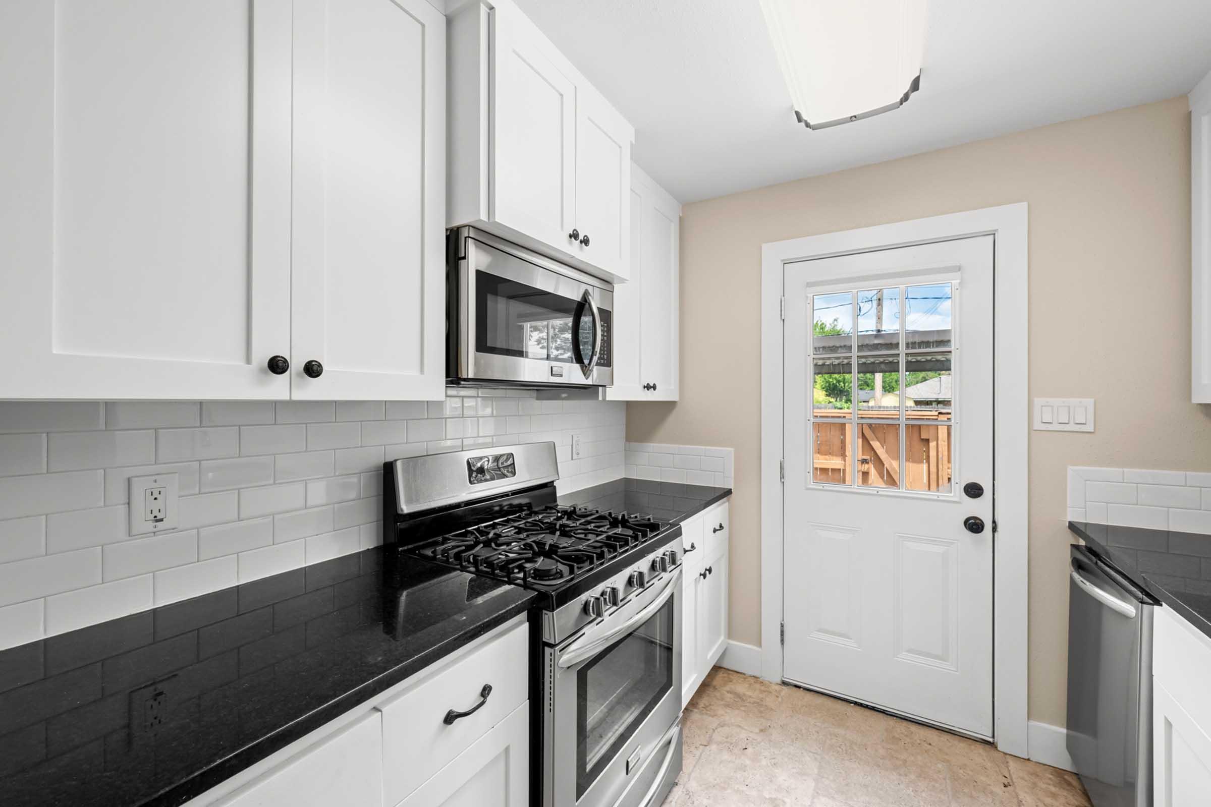 A modern kitchen featuring white cabinetry, a stainless steel microwave, and a gas stove with a sleek black countertop. There's a door leading outside, with light-colored walls and a window above the sink, creating a bright and inviting atmosphere.