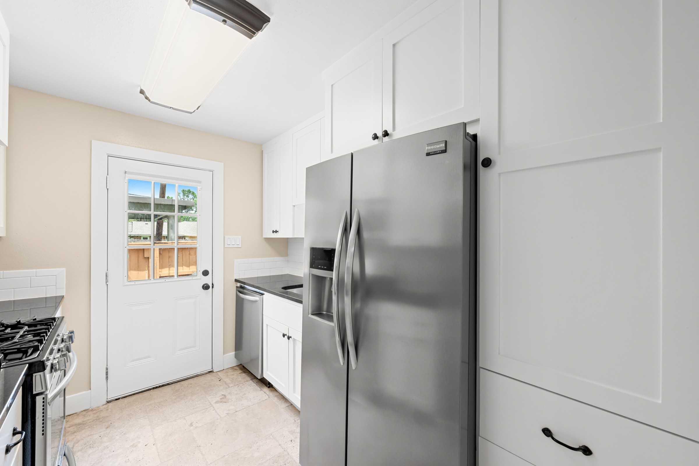 Modern kitchen featuring stainless steel appliances, including a refrigerator and stove. The white cabinetry contrasts with light-colored walls and tile flooring. A door leads outside, allowing natural light to brighten the space. The layout is functional and inviting, suitable for everyday cooking and entertaining.