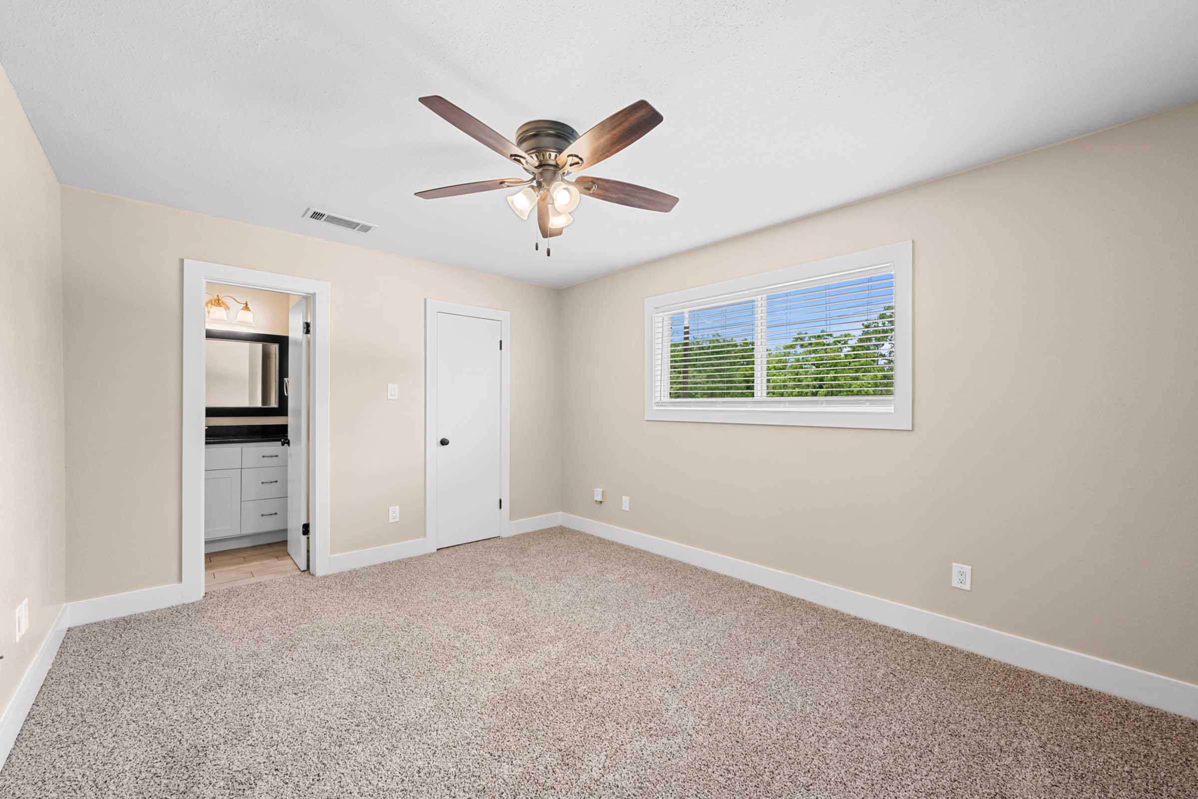 A spacious, well-lit room featuring a light-colored wall, a ceiling fan with wooden blades, and a plush carpeted floor. A window with blinds allows natural light, and a door leads to a bathroom. The room has a clean and modern aesthetic, suitable for various uses.