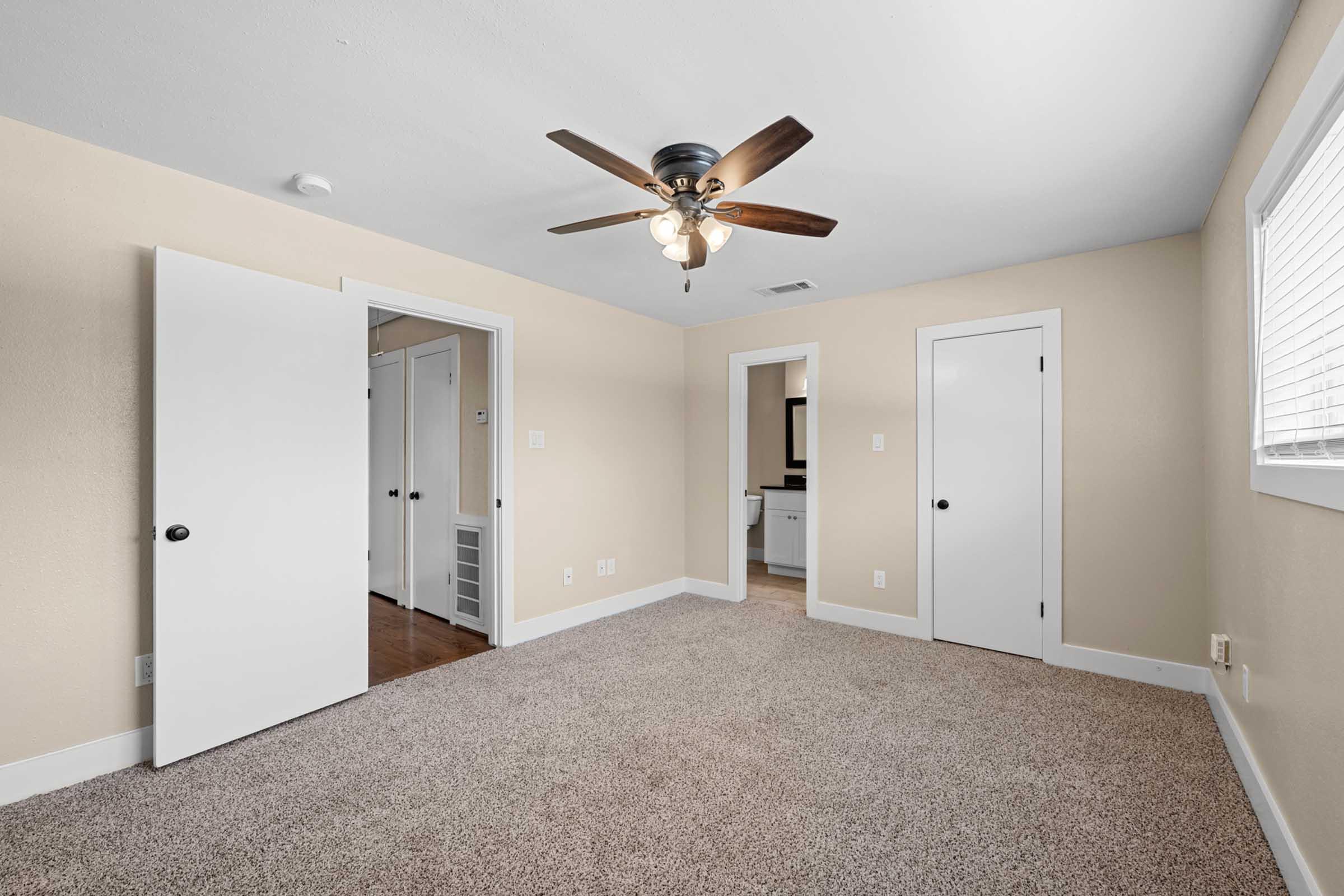 A well-lit room featuring beige walls and carpeted flooring. There is a ceiling fan with wooden blades, and two doors leading to adjacent spaces. A window allows natural light to enter, enhancing the airy feel of the space. The overall atmosphere is clean and inviting, suitable for various uses.