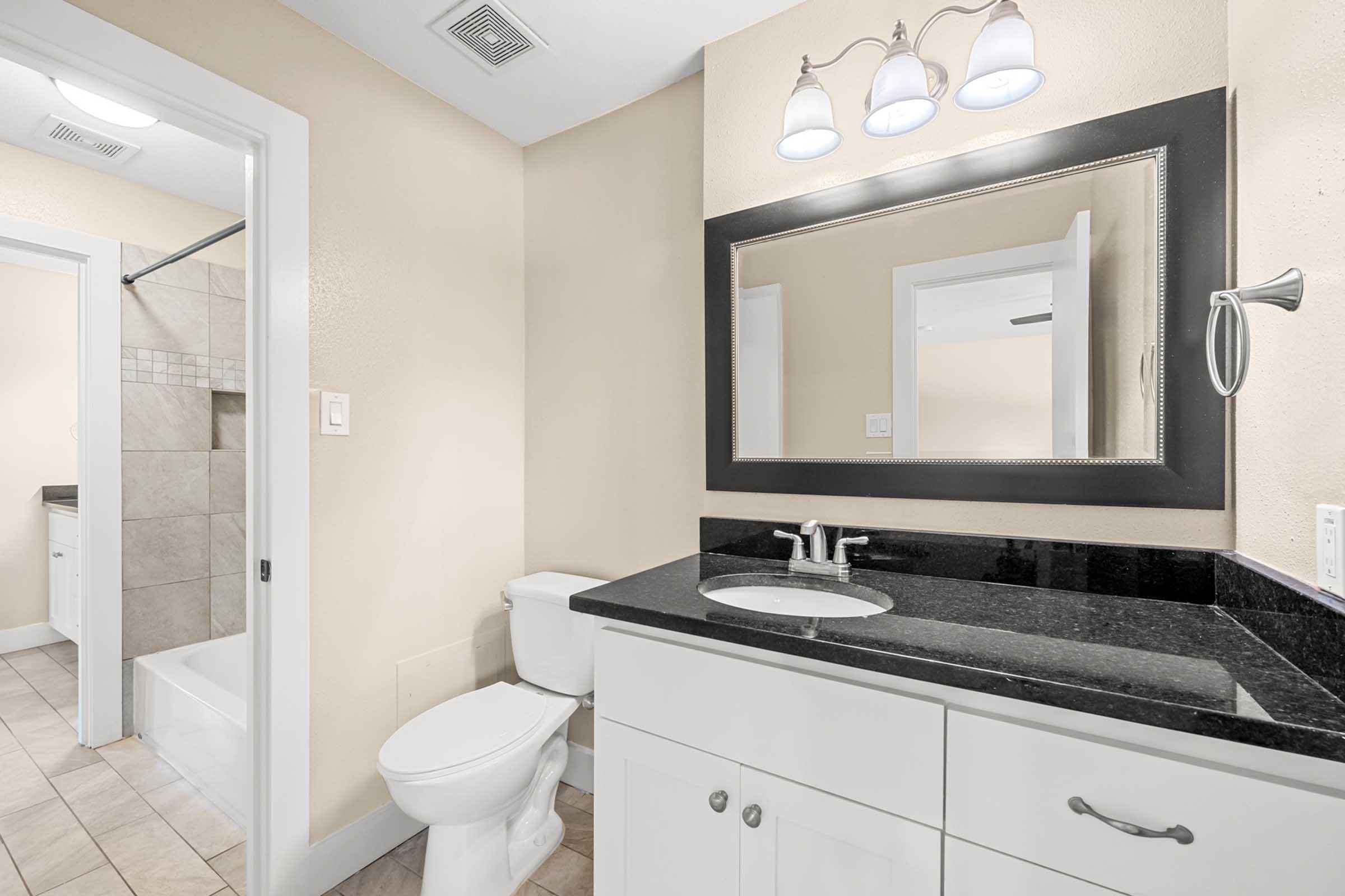 A bathroom featuring a white sink with a dark countertop, a large mirror with a dark frame, a toilet, and tiled flooring. The room has light beige walls and a shower area partially visible in the background. Three light fixtures are mounted above the mirror.