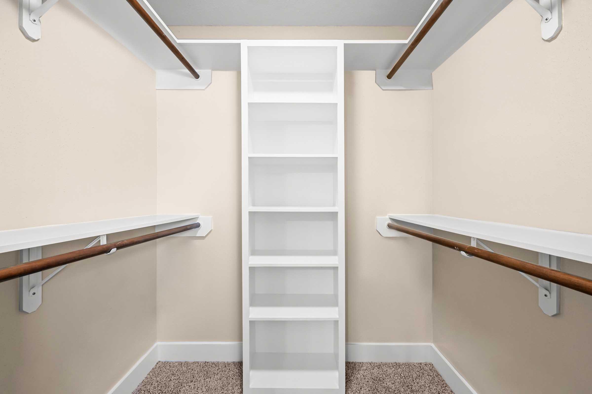Empty walk-in closet featuring two wooden hanging rods on either side and a central white shelving unit. The walls are painted a light color, and the floor is covered with carpet. Good lighting enhances the spacious feel of the area.