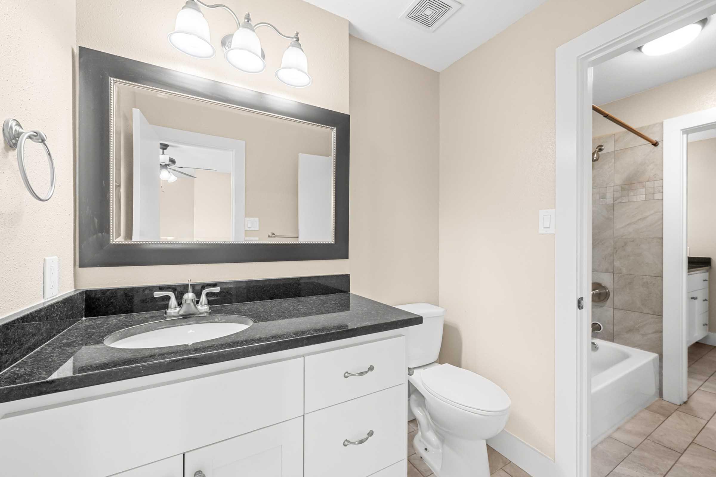 A modern bathroom featuring a black granite countertop with a sink and mirror above, a wall-mounted light fixture, a toilet, and a glass shower enclosure. The walls are painted a light beige, and there is tiled flooring. A doorway leads to another room in the background.
