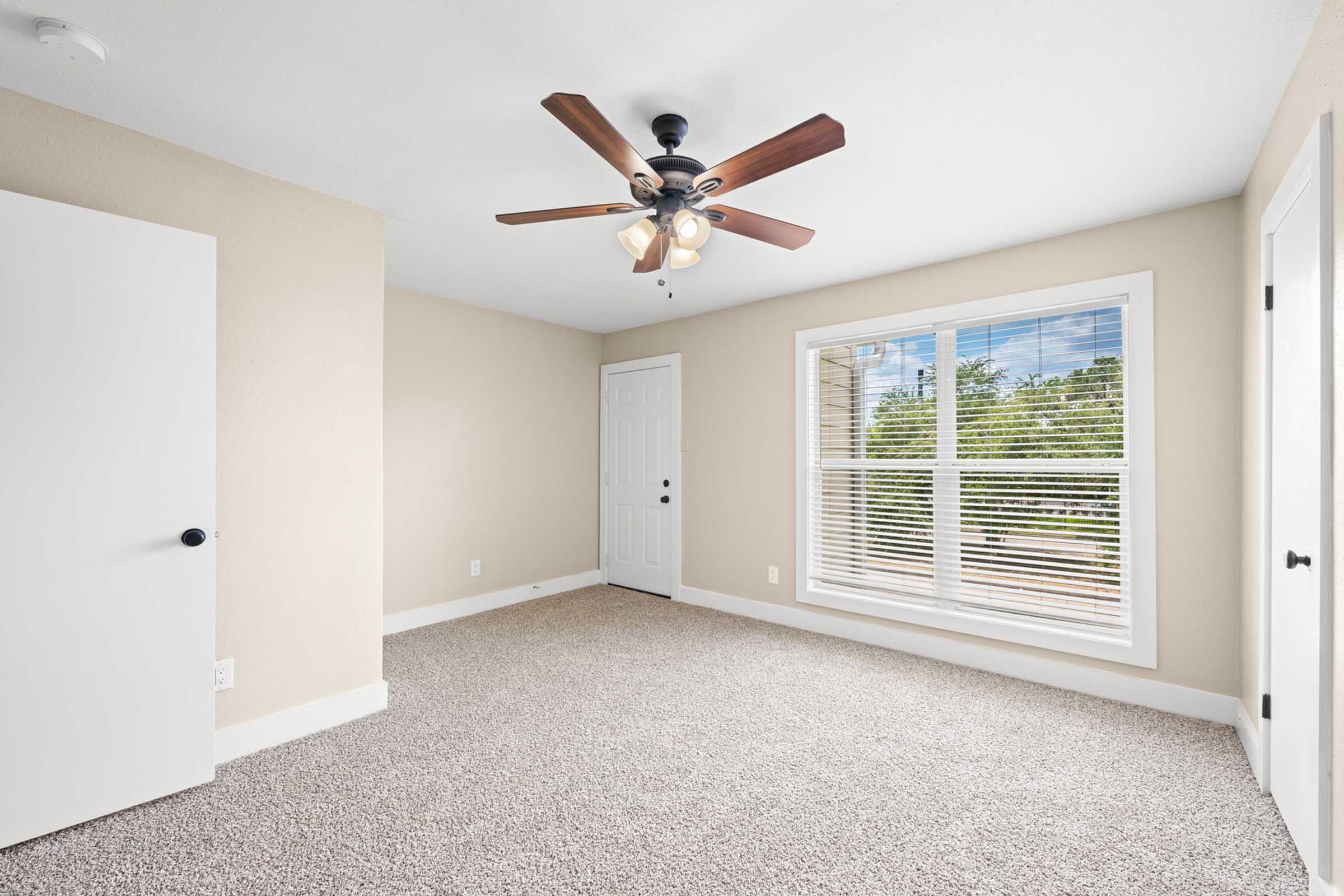 A light and airy room with beige walls and a ceiling fan featuring wooden blades. The room has a plush carpet, a large window with white blinds letting in natural light, and a solid white door. It's empty, suggesting it's ready for furnishings or personal touches.