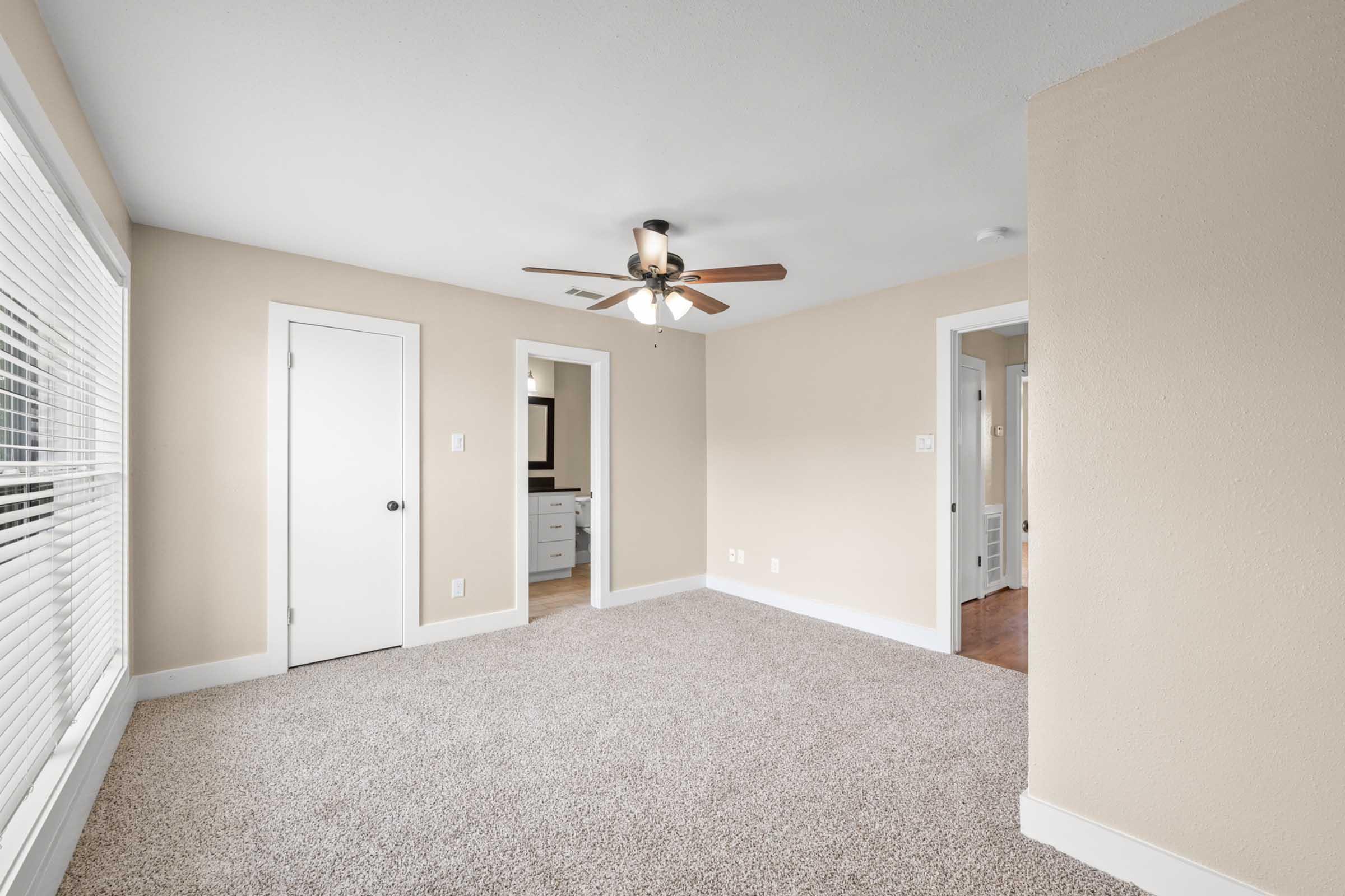A neatly arranged room with light-colored walls and carpet. There's a ceiling fan, a window with blinds allowing natural light, and a door leading to another area. The space appears well-maintained and bright, suitable for various uses like a bedroom or office.