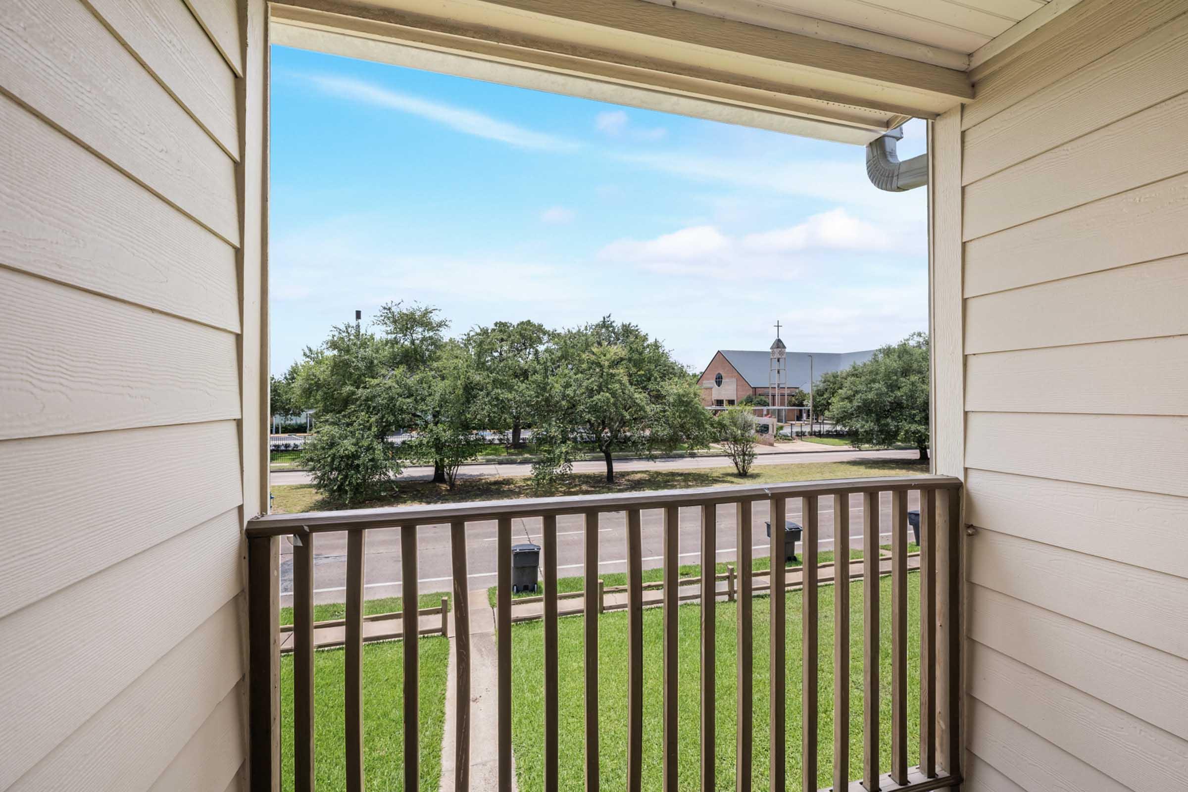 View from a balcony showing a wooden railing. Below, green grass and a tree line are visible, with a wide street in the foreground. In the background, there are buildings and a cloudy blue sky.