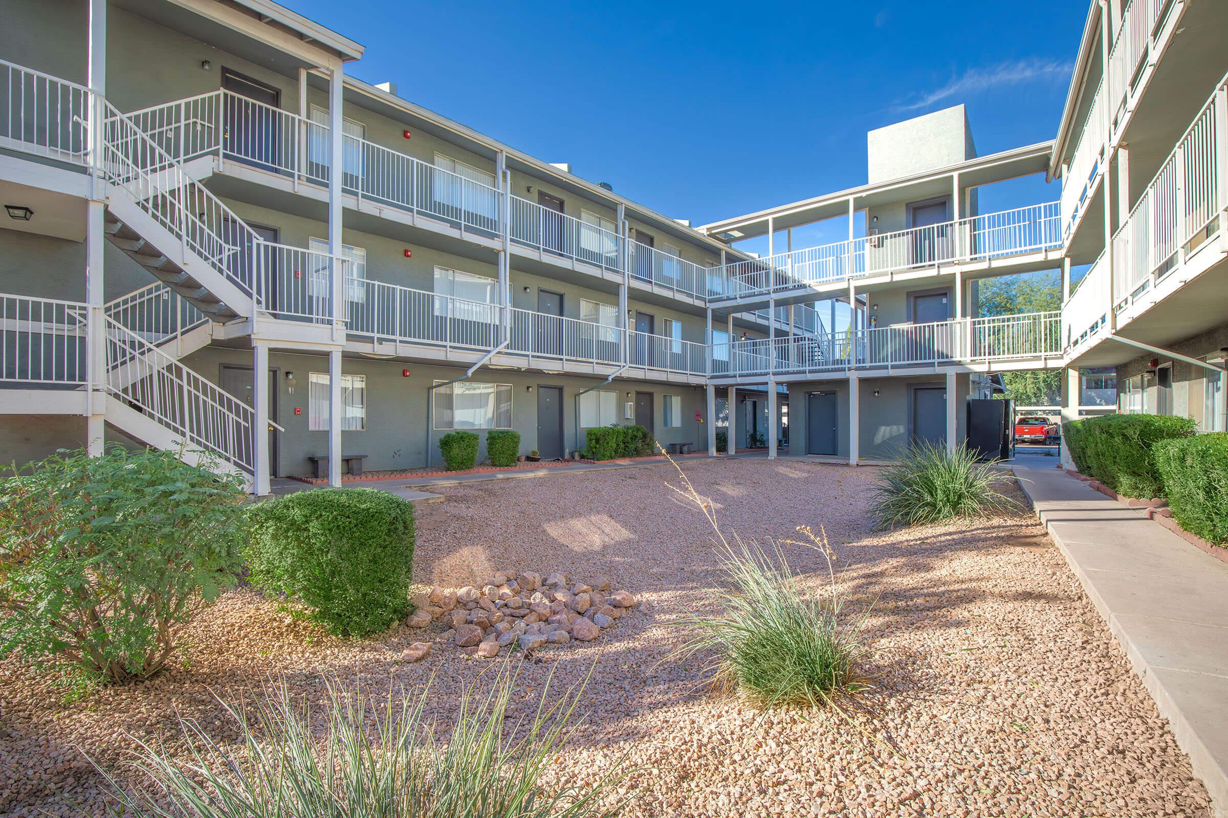 A view of a multi-story apartment complex with a courtyard featuring desert landscaping, including gravel, shrubs, and small rocks. The building has white railings and balconies, with clear blue skies above.