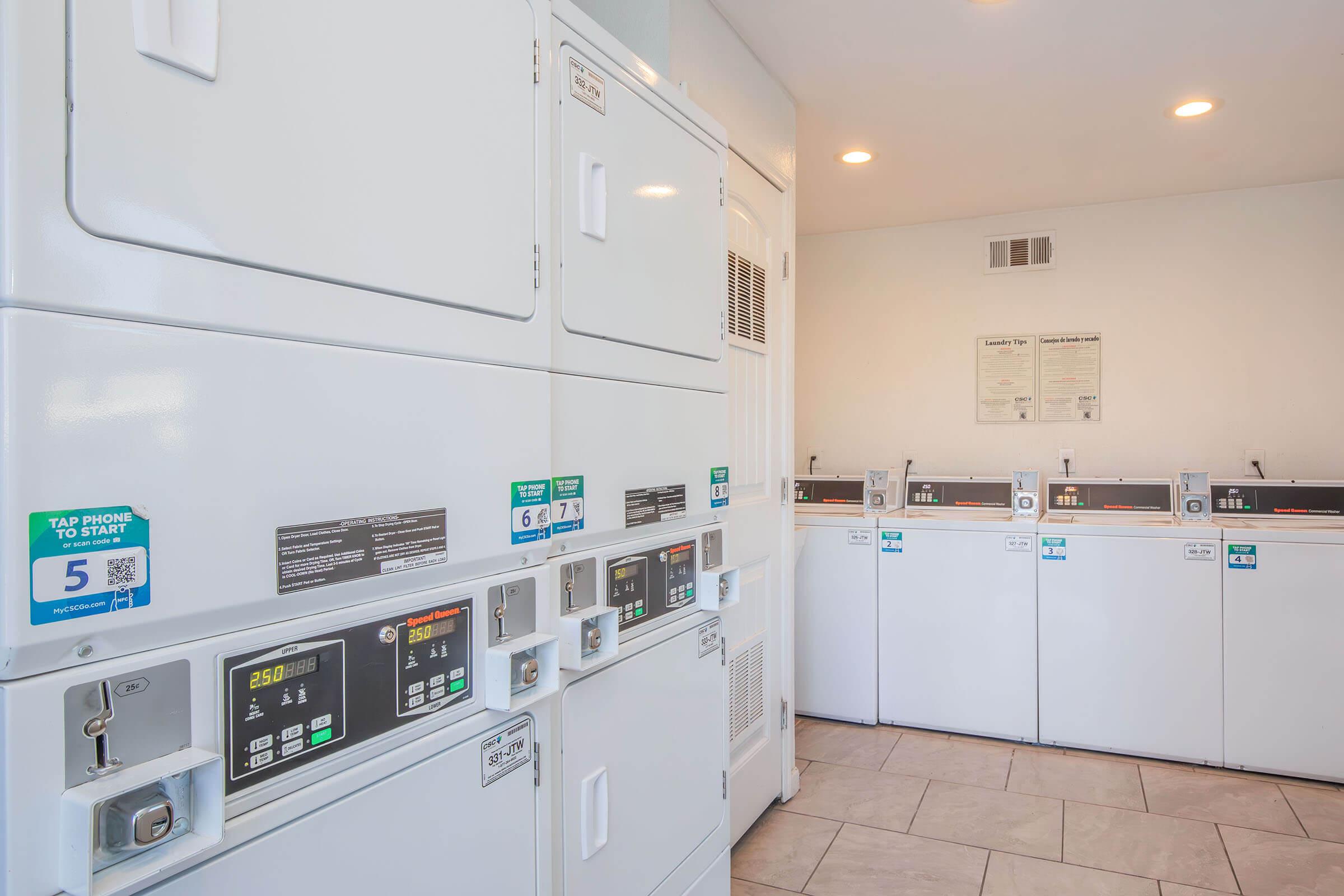 A modern laundromat featuring multiple front-loading washing machines and dryers. The appliances are neatly arranged against a wall, equipped with digital control panels displaying instructions and pricing. The light, clean interior has tiled flooring and bright lighting.