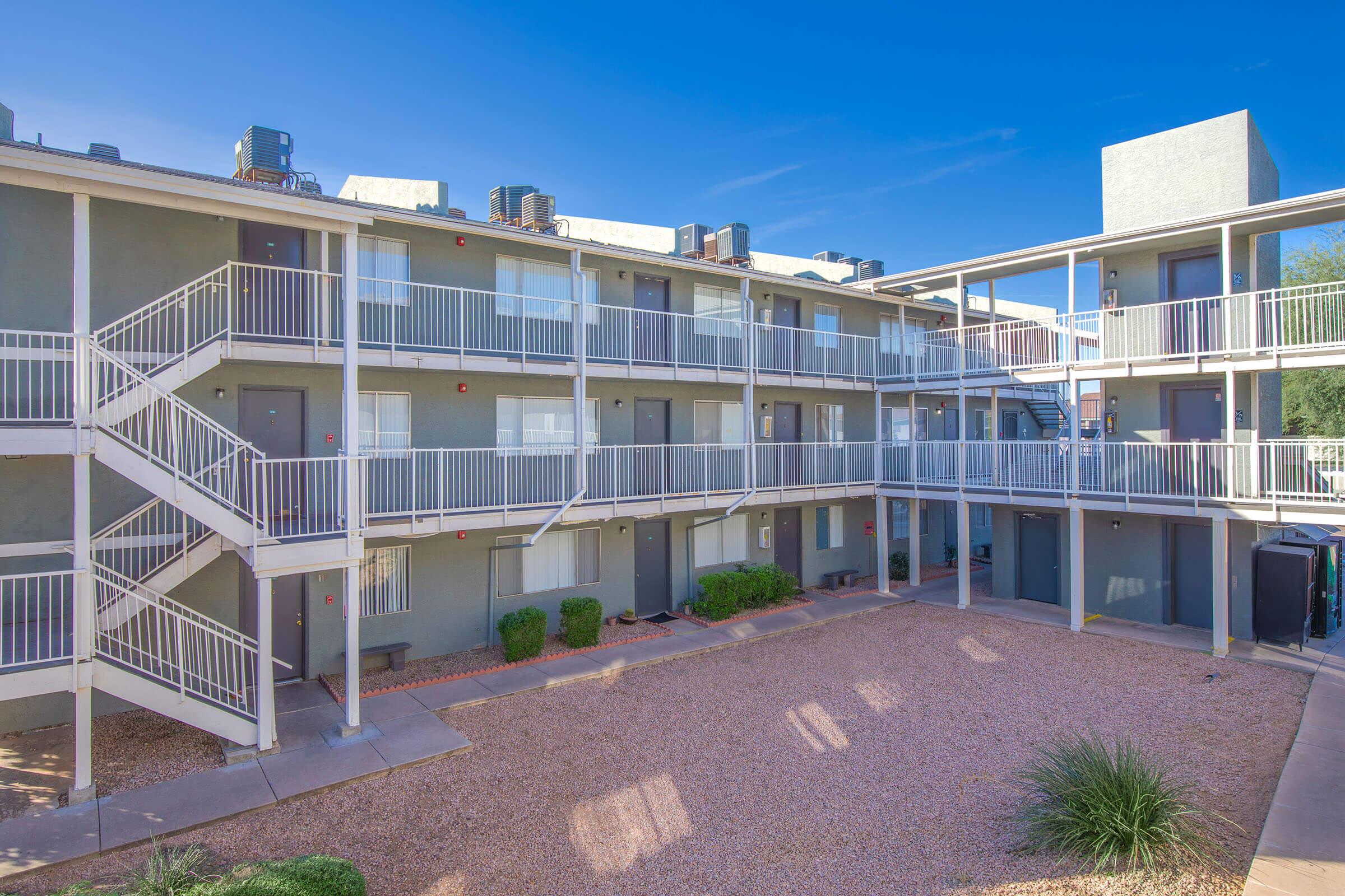 A view of a multi-story apartment building with a central courtyard. The two-level structure features white railings on the balconies and several air conditioning units on the roof. There are stone pathways, landscaped areas, and a clear blue sky in the background.
