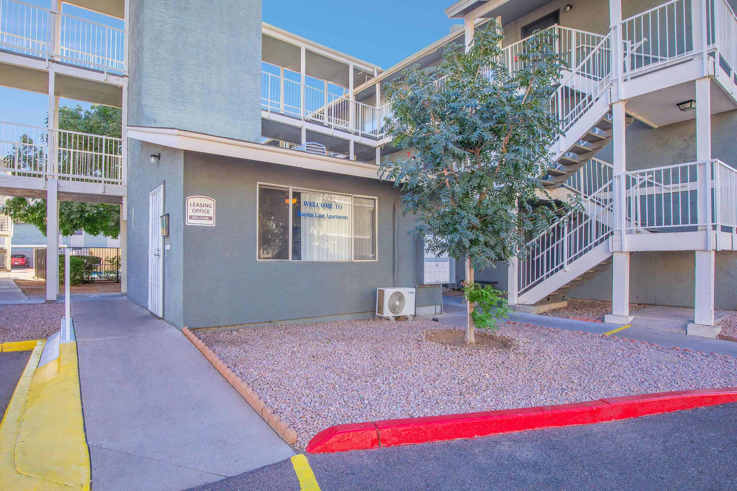 Exterior view of an apartment building featuring a staircase, landscaped area with a small tree, and an air conditioning unit. A sign indicates a leasing office nearby. The building has a modern design with a light grey color and white railings. The pathway is bordered with a red curb.