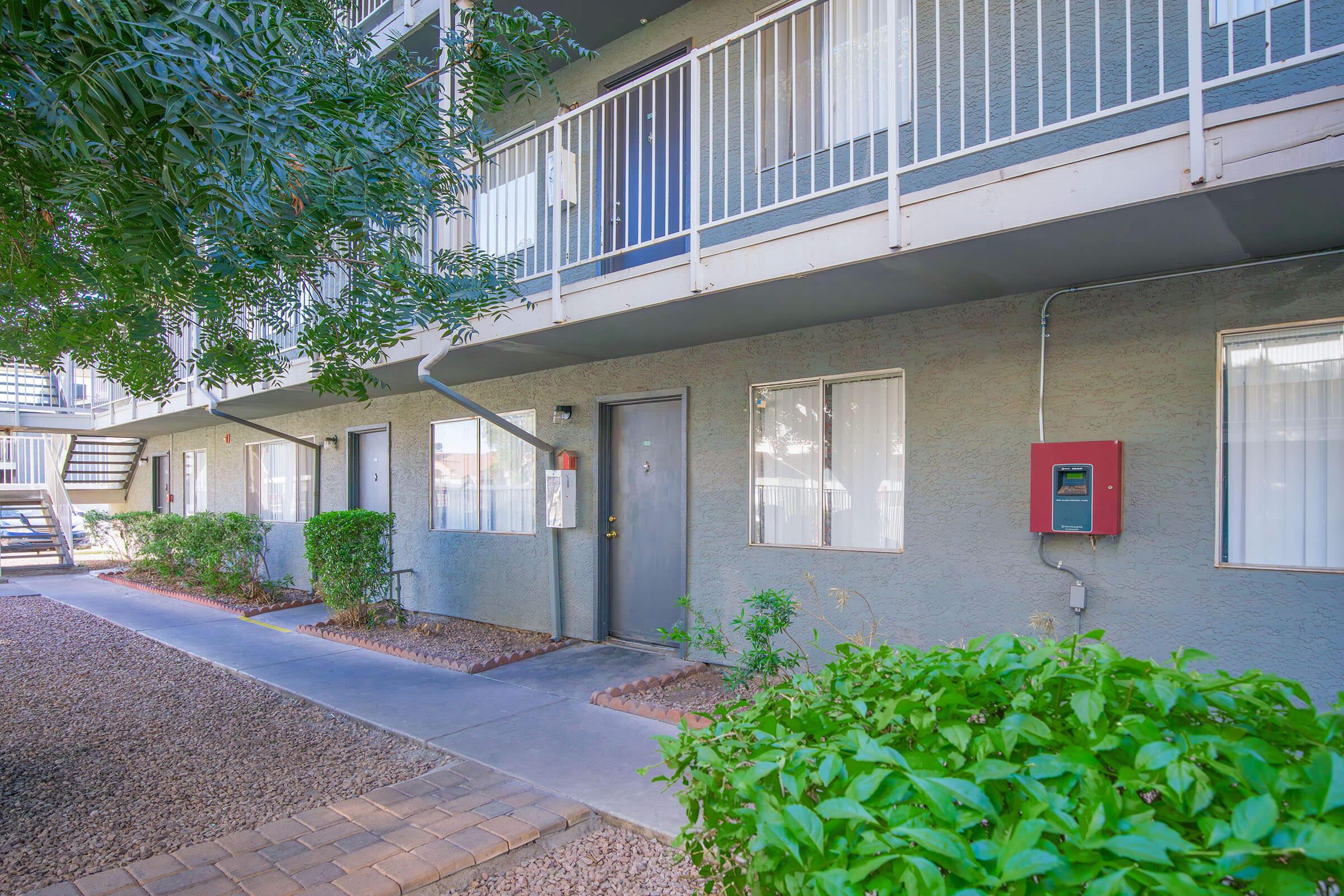 A view of an apartment building exterior with a gray facade. There are multiple windows with white curtains and a central front door. Surrounding the building are landscaped areas featuring small bushes and a pathway made of stone pavers, along with a staircase leading to the upper floor.