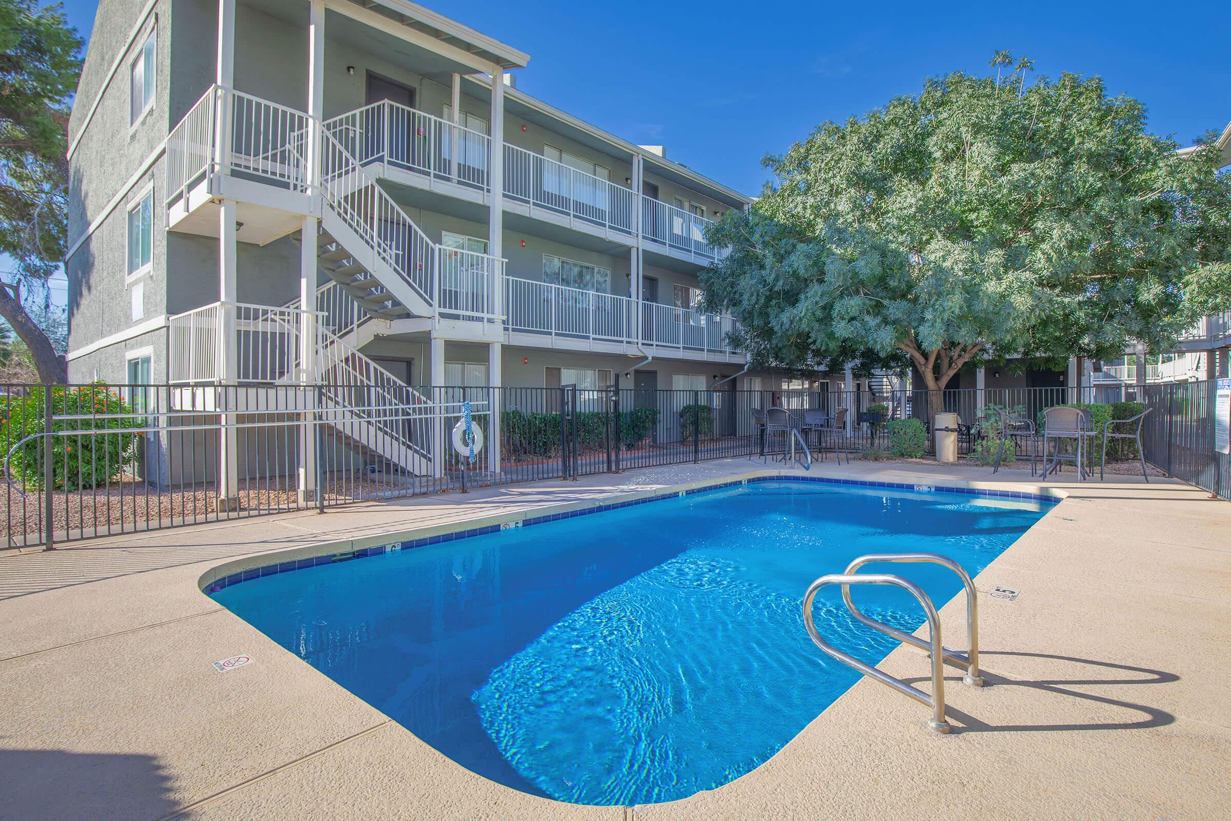 A clean, inviting outdoor pool area with crystal-clear water, surrounded by a gated fence. In the background, a three-story apartment building with a stairway and balconies can be seen, alongside a lush green tree providing shade. The sky is clear and blue, creating a pleasant atmosphere.