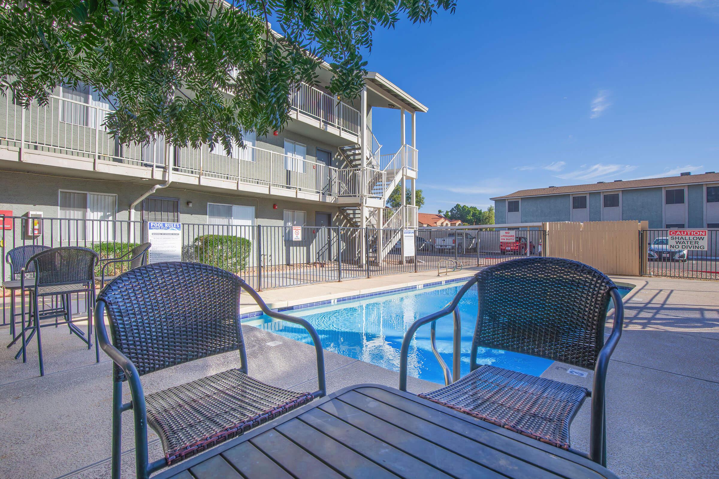 A sunny view of a pool area surrounded by apartment buildings. Two black wicker chairs and a small table are in the foreground, overlooking the clear blue water of the pool. The scene includes trees and a fenced area, contributing to a relaxing outdoor atmosphere.