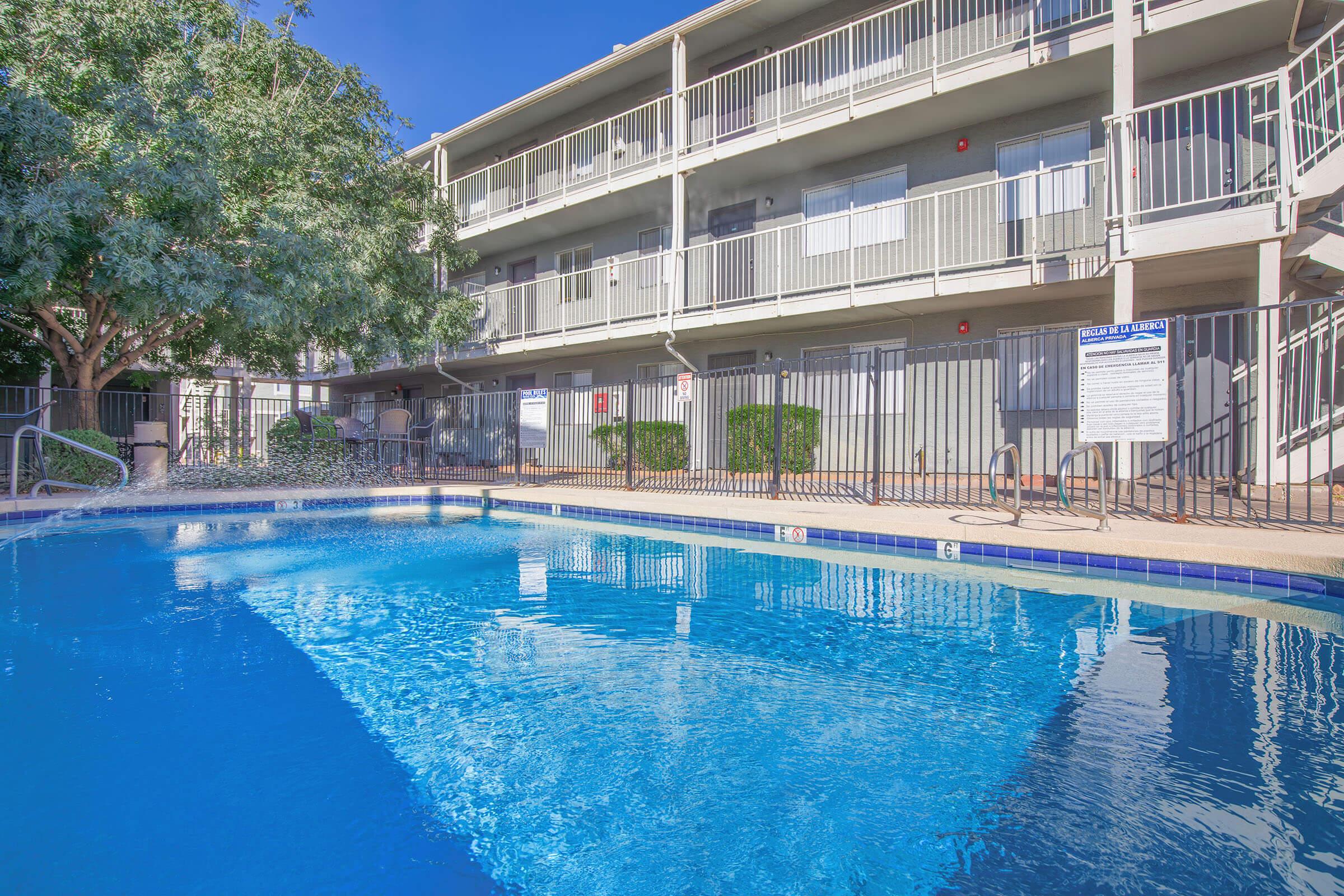 A clean swimming pool surrounded by a railing, with a multi-story apartment building in the background. The building features balconies and large windows. Lush greenery can be seen nearby, and the sky is clear and blue, reflecting on the surface of the water.
