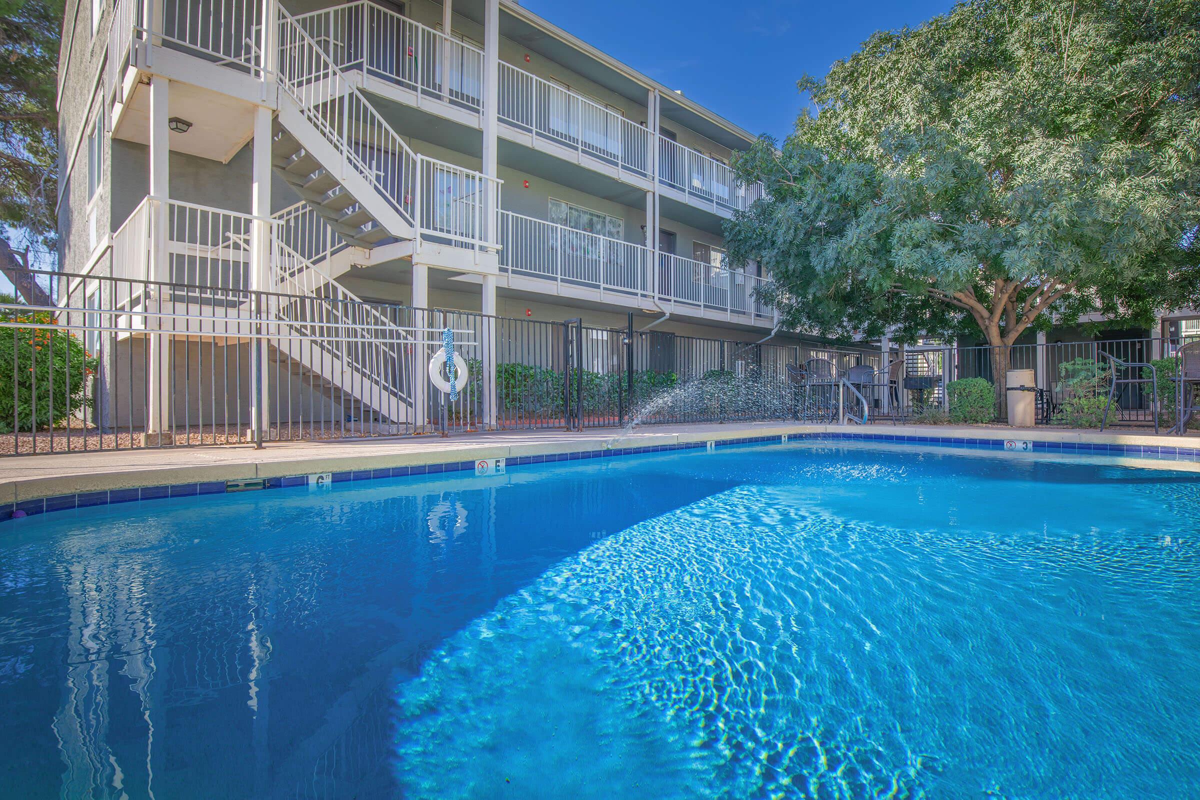 A clear blue swimming pool with a fountain in the foreground, surrounded by a gated area. In the background, a multi-story building with a staircase and greenery. The sky is blue with a few clouds, creating a bright, inviting atmosphere.