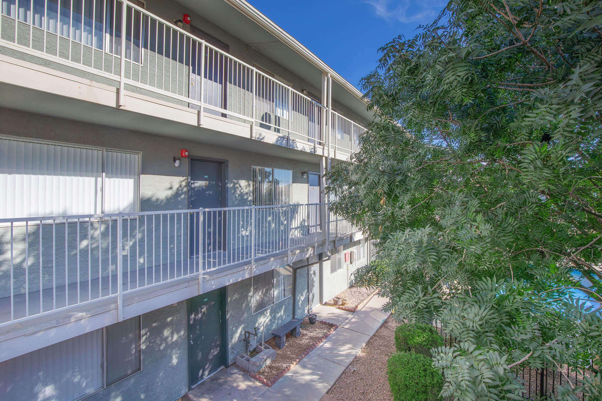 Three-story apartment building with balconies, light-colored exterior, and stairway access. Surrounding greenery includes a tree and shrubbery, while the sky is clear and blue. The pathway leads through landscaped areas, emphasizing a well-maintained outdoor space.