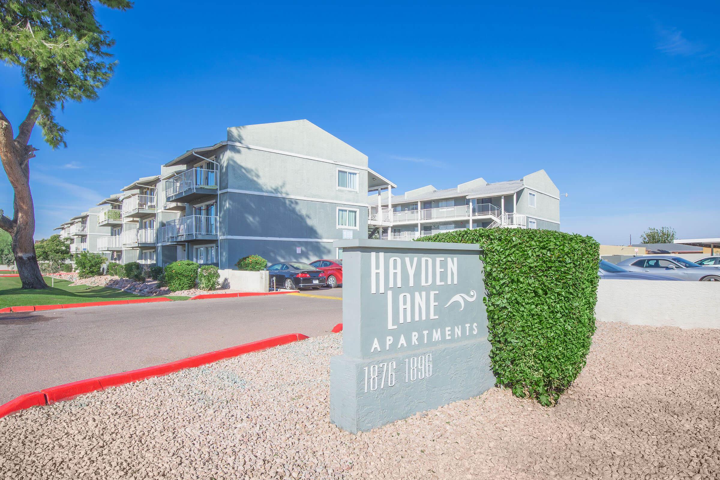 A view of Hayden Lane Apartments, featuring a multi-story building with a light gray exterior and balconies. The sign in front prominently displays "Hayden Lane Apartments" along with the address range of 1876-1986. Surrounding the property are landscaped areas with gravel and trees under a clear blue sky.