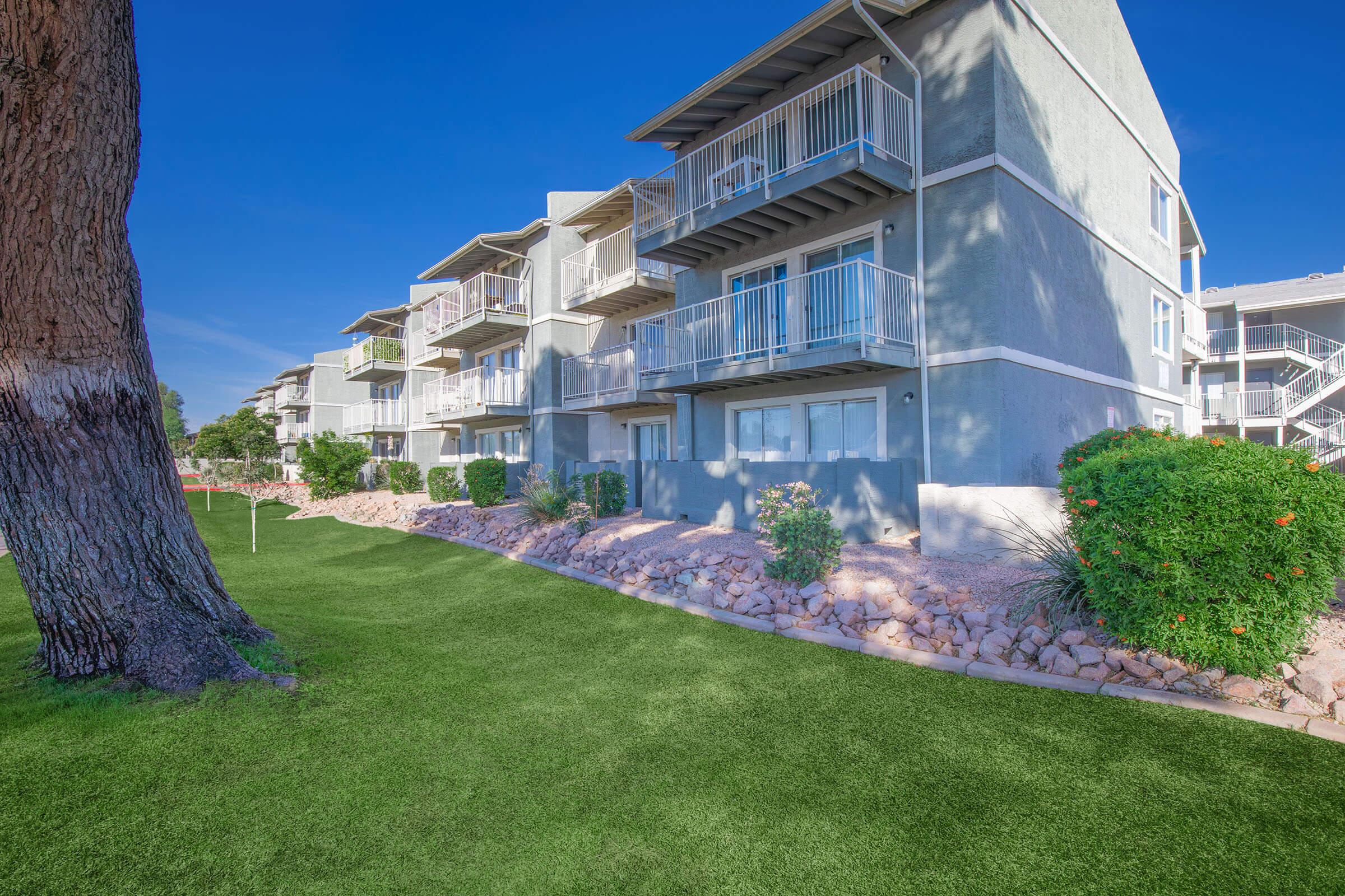 A modern apartment building with several balconies and large windows. The structure features light gray walls and is surrounded by a well-maintained landscaping with plants and a green lawn, along with rocky elements in the garden area. Bright blue sky in the background.