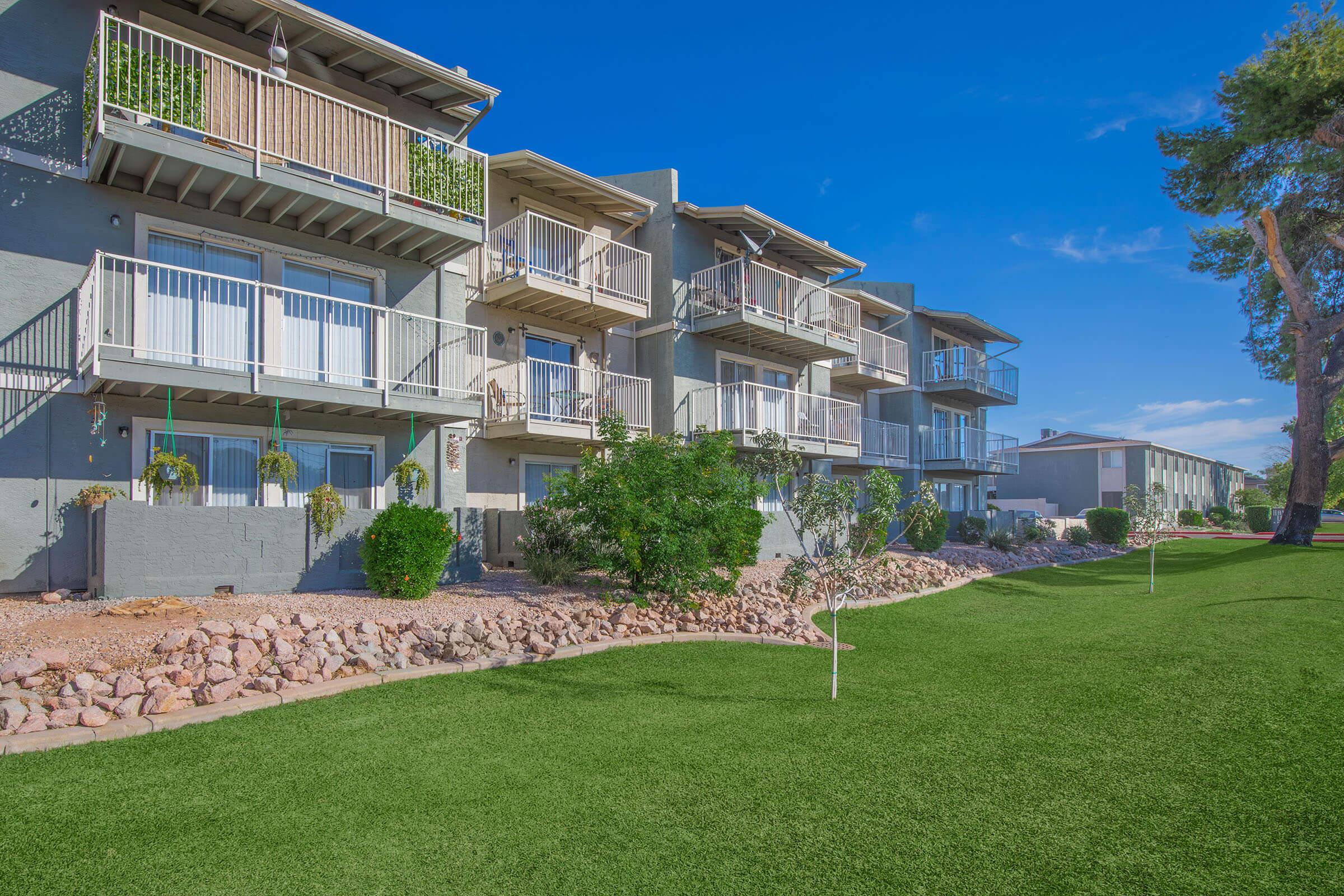 Apartment complex featuring multiple buildings with balconies, surrounded by green grass and landscaped rocks. The scene is set against a clear blue sky, showcasing a well-maintained outdoor area ideal for residents.