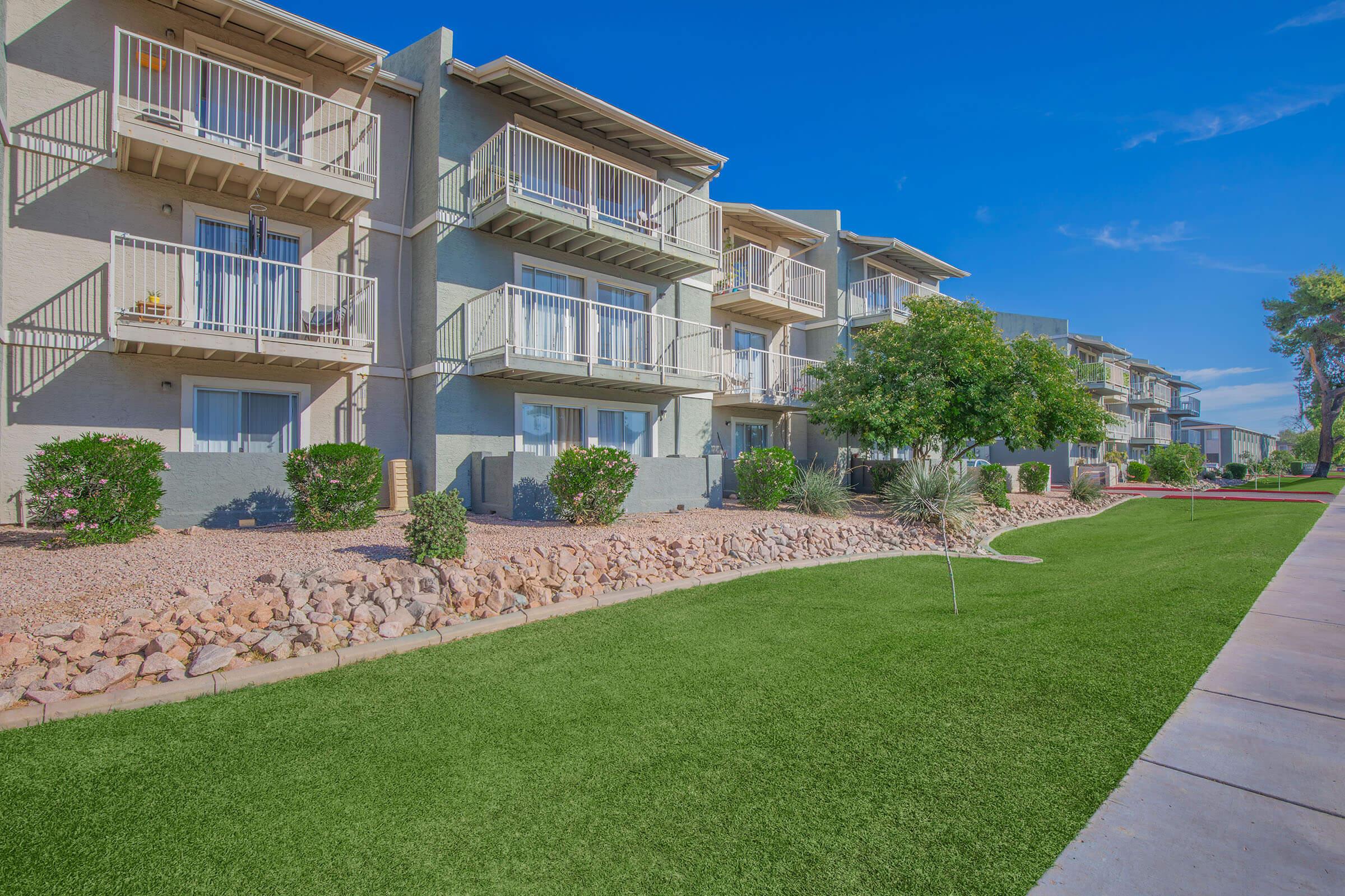 A row of apartment buildings with balconies, set against a bright blue sky. The ground features a well-maintained grassy area bordered by rocks and shrubs. Nearby, trees provide shade, creating a pleasant outdoor atmosphere.